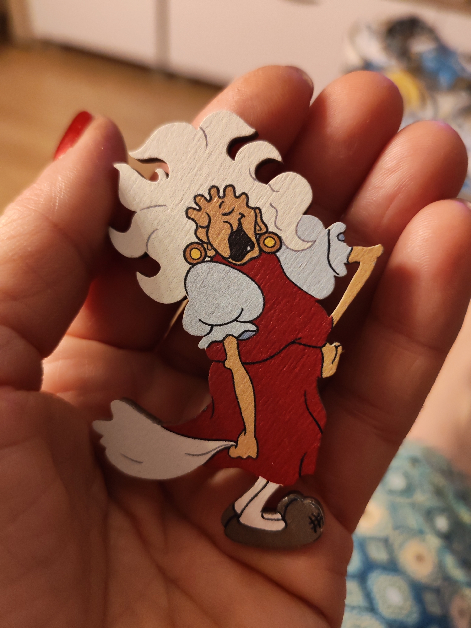 A person is holding a wooden cartoon depiction of a woman. The woman has a large white wig, is wearing large gold earrings, a red dress with light blue sleeves, and brown shoes. She is yelling with her mouth open and is holding a walking stick. The person's hand is pale with a red fingernail. The background is blurred, showing a wooden surface and a patterned blue cloth. There is no clear indication of Chaikovsky, Russia.
