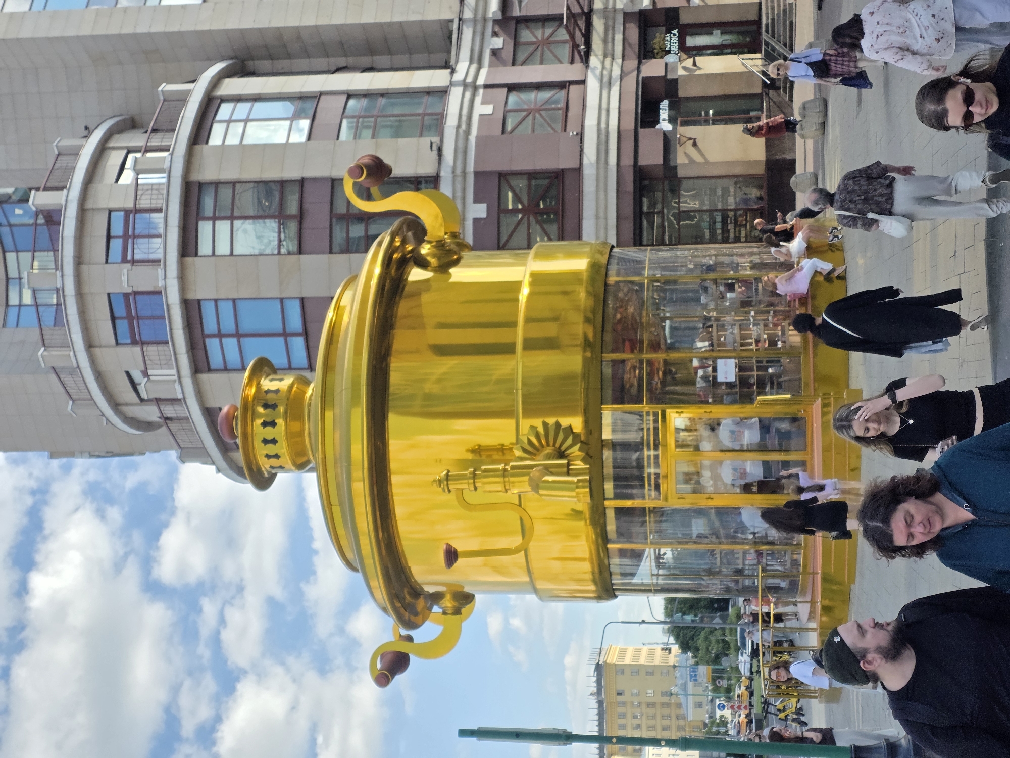 This media file features a large, golden samovar sculpture prominently in the foreground, set against a backdrop of a modern building with numerous windows. The image appears to be taken during the daytime under a sky with scattered clouds. Several people are visible in the scene, some walking along a paved walkway and others gathered near the sculpture. Their attire suggests a casual, possibly warm weather setting. Reflections in the glass panels of the building and the samovar itself offer glimpses of other individuals and the surrounding environment. One section of the building has text that reads "NATURA SIBERICA" and another section has "ДЫМЕЛЬ". The overall mood seems relaxed, with people going about their day in an urban setting.