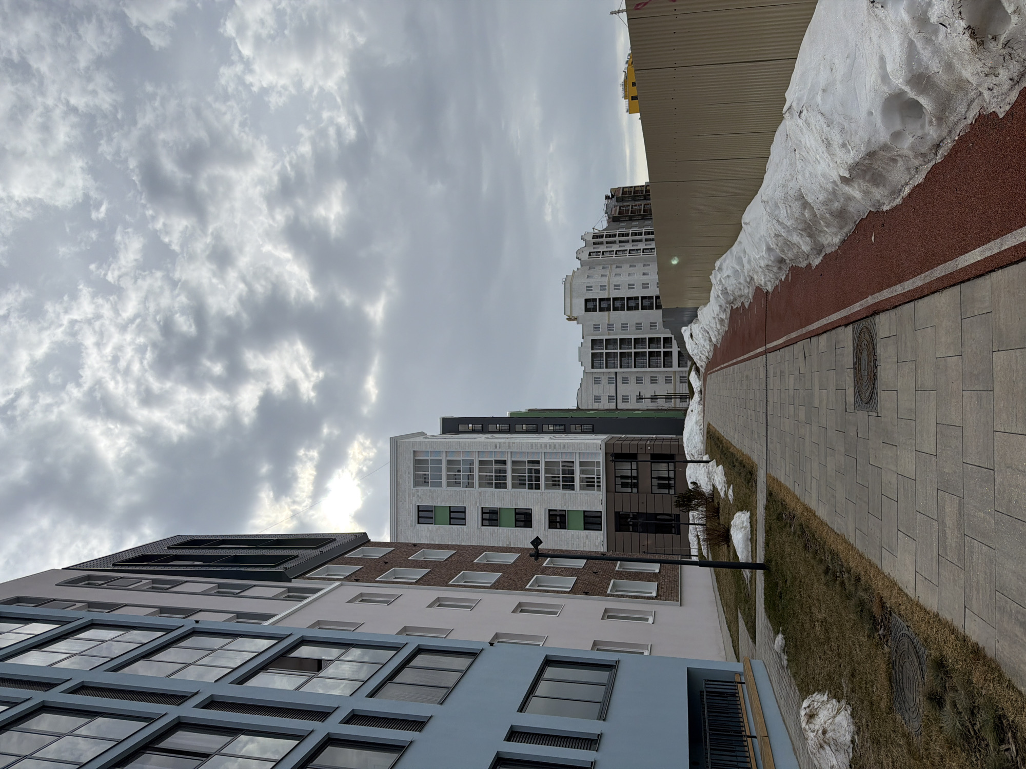 The image depicts a street scene in what appears to be a residential area in Pervomaysky, Russia, on a cloudy day. Several modern apartment buildings of varying heights and colors (blue, beige, white, brown) line the street. The buildings have a variety of window styles and architectural features. A pedestrian walkway paved with gray bricks runs along the right side of the frame. A strip of grass separates the walkway from a red surface covered in patches of snow. A large pile of snow sits atop the red surface, adjacent to a building. Streetlights are visible along the walkway. The sky is overcast with grey clouds. The image is taken from a low angle, looking upwards towards the buildings and sky.