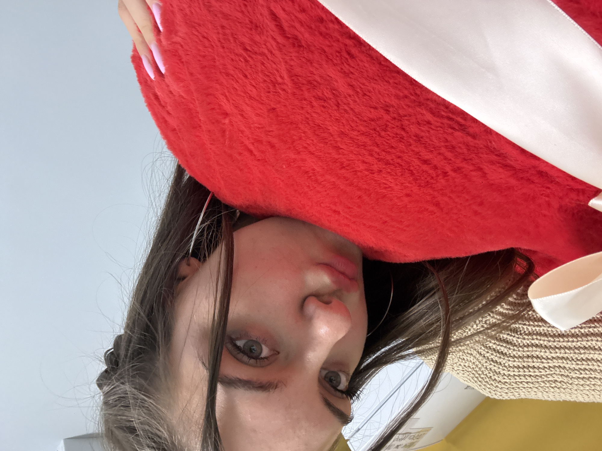A young woman with long brown hair and pink fingernails is holding a large, red, heart-shaped plush object with a white ribbon. The image is taken from a low angle, looking up at the woman's face. She has light skin, brown eyes, and a slightly pouting expression. The background is a plain, light-colored wall. The location is not discernible from the provided image.