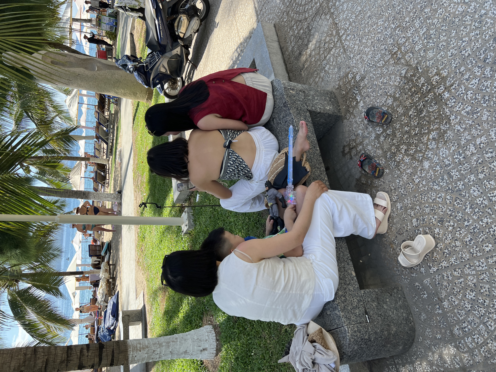 In Nha Trang, Vietnam, a group of people are relaxing at a beachside resort. In the foreground, three women and a young boy sit on a stone bench. Two women are facing away from the camera, one wearing a white top and white pants, and the other in a red top and white pants. The third woman, also in white, is seated with her arm around the boy. A fourth person, the boy, is looking down and playing with toys. Scattered on the ground are various sandals and a straw hat.
In the background, palm trees line a sandy beach dotted with lounge chairs and umbrellas. People in swimwear can be seen walking and lounging near the ocean. The water is calm, and a clear blue sky stretches overhead. A parked motorcycle is visible on the right side of the frame. The overall scene suggests a leisurely day at a tropical vacation destination.