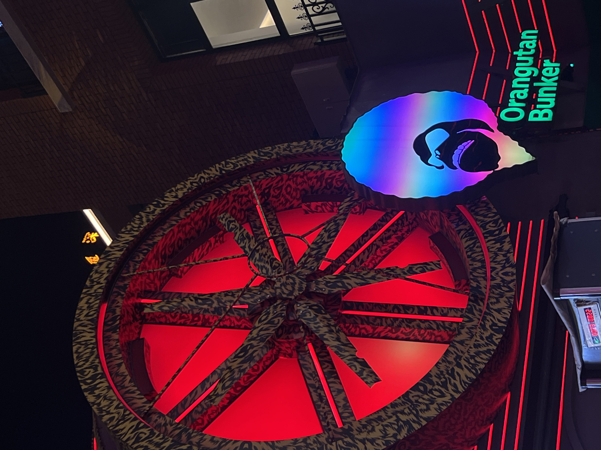 The image shows an elaborate neon sign and architectural details in Futian District, China. The sign features a large, circular structure with a red neon-lit interior and a tiger-striped patterned exterior. Radiating from the center of the circle are multiple tiger-striped bars that resemble spokes. Above the circle is a smaller, cloud-shaped sign with a depiction of an orangutan's head, illuminated in rainbow colors. Next to the orangutan sign, green neon lettering reads "Orangutan Bunker." Vertical red neon lines run along a dark-colored wall near the right edge of the image, and a metallic object is also attached to the same wall. Further above, a window with a dark frame and ornate metalwork is visible against a brick wall. The overall scene has a dark and vibrant, urban nightlife atmosphere.