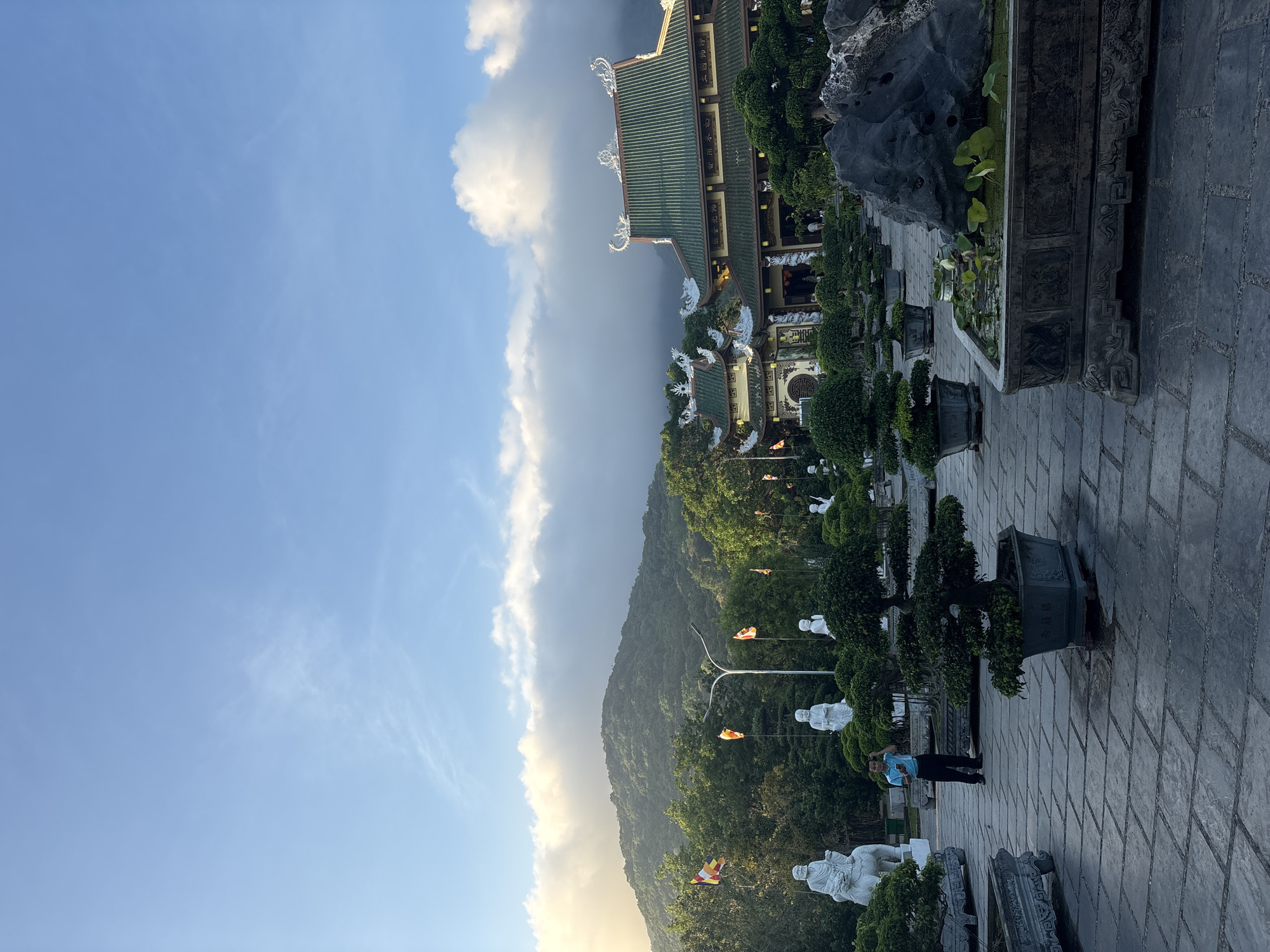 The image shows a view within the grounds of the Linh Ứng Temple in Đà Nẵng, Vietnam. The perspective is from a paved walkway, oriented with the sky and mountain to the left and a section of the temple buildings to the right.
The left side of the image is dominated by the sky, partly cloudy with a blue hue. Below the sky is a mountain, heavily forested. Along the slope of the mountain, a series of white statues are visible, possibly figures of worship, as well as streetlights.
On the right side, there is a temple structure featuring traditional green tile roofs and ornamental details. Below the temple building, the walkway is lined with trees, manicured shrubbery, and several large stone planters and more statues.
In the lower right of the frame, a person wearing a blue shirt is present.
The general environment suggests a serene, elevated location overlooking the surrounding landscape.