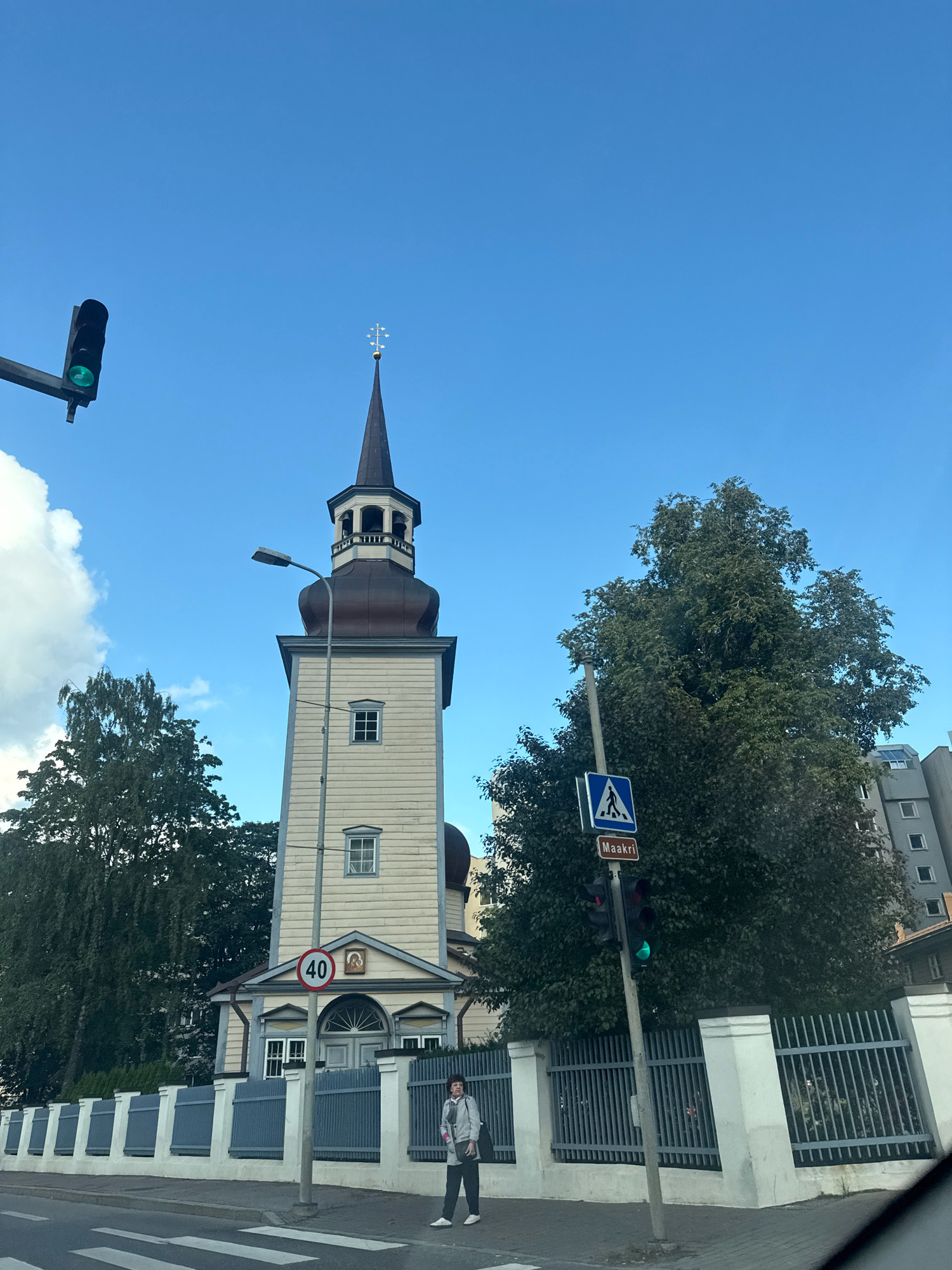 The image depicts a street scene in Tallinn, Estonia, featuring the Orthodox Church of St. Simeon and St. Anna. The church's light-colored wooden tower with a dark spire is prominent against a clear blue sky with some scattered clouds. A traffic light displaying green is visible on the left, and another traffic light, also green, is on the right. A pedestrian crossing sign with "Maakri" written below it is also present near the traffic light. A speed limit sign indicating 40 is attached to a pole in front of the church entrance. A person dressed in casual clothing is standing on the sidewalk next to a metal fence. Trees are visible in the background, along with a modern apartment building. The foreground shows a crosswalk on a paved street.