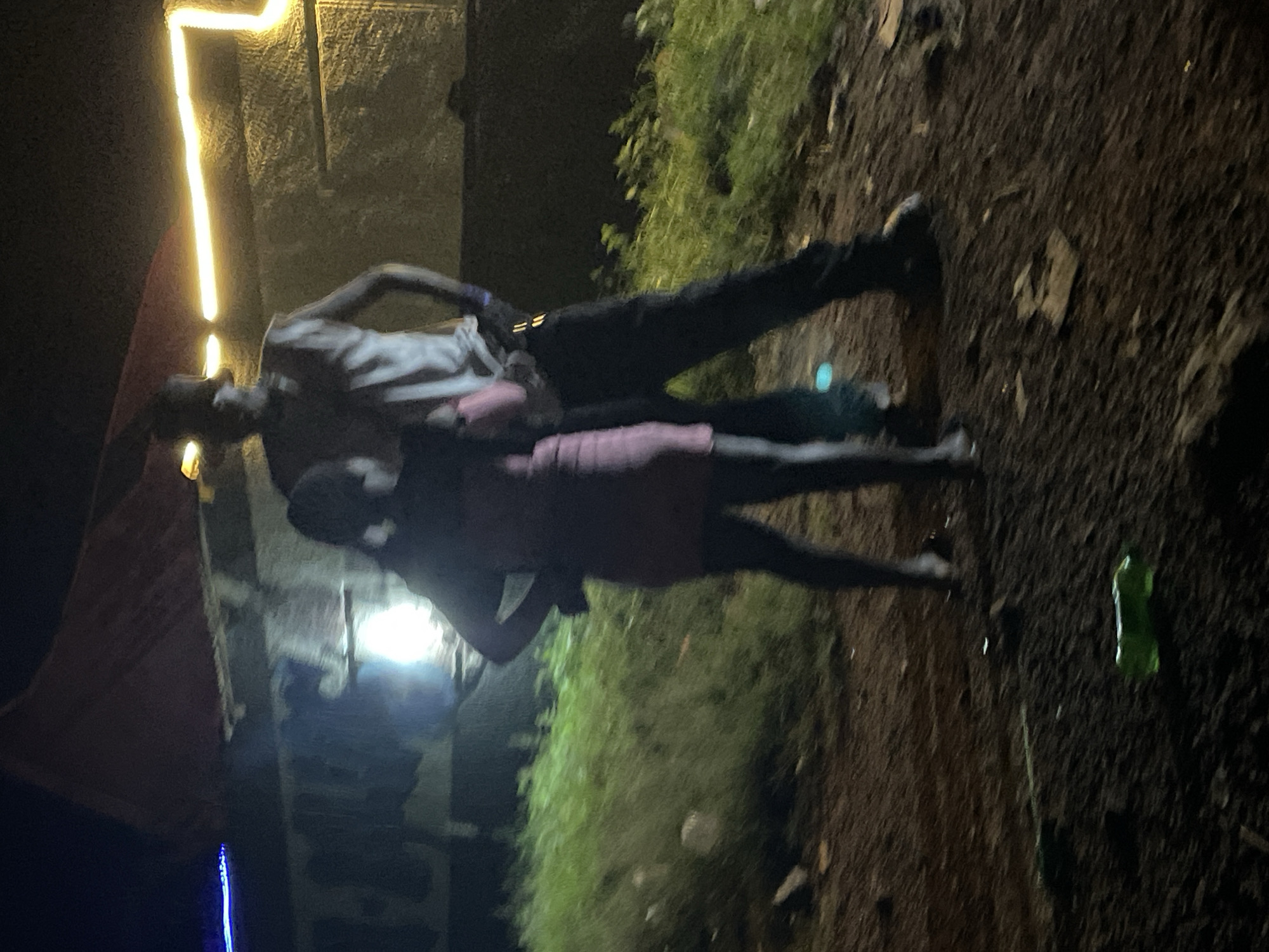 Two individuals are standing in an outdoor environment at night. The person on the left is wearing a pink dress and dark leggings, with their hands on their hips. The person on the right is wearing a light-colored sleeveless shirt, dark pants, and a cap. They appear to be standing close together. Behind them, a structure with a red roof and a wall illuminated by yellow lights is visible. The ground is rough and appears to be dirt with some grass. A green plastic bottle is on the ground in the foreground. The scene is dimly lit, suggesting it is a rural or unlit urban area, possibly on the outskirts of Durumi, Nigeria.