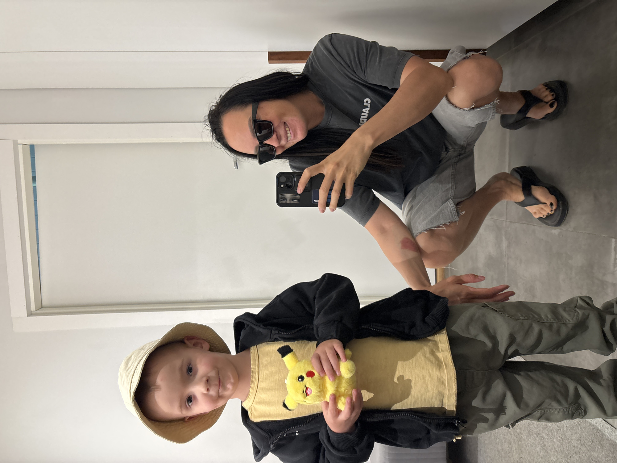 A person with long dark hair, wearing sunglasses, a gray t-shirt, and cut-off shorts, squats on a gray floor and holds a smartphone in their right hand. The person is smiling and appears to be taking a photo. In the foreground is a young child wearing a tan bucket hat, a yellow t-shirt with a Pikachu graphic, a black jacket, and green pants. The child is holding a Pikachu plush toy. A white wall is visible to the left, with what appears to be a window or door frame. Behind the person is another section of the white wall and a wooden bar. The location cannot be determined from the image but the city of Chaikovsky, Russia, is mentioned as a request.