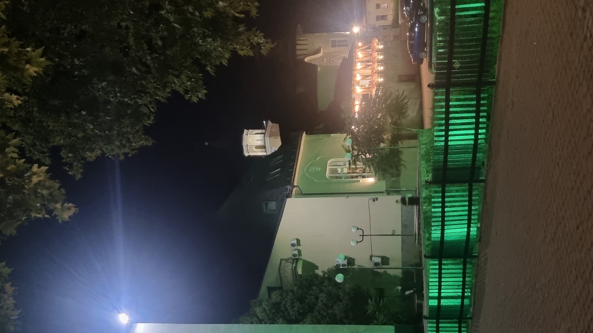 This vertical, nighttime photograph captures a street scene in Berehove, Ukraine, featuring a building with light-colored walls and green accents, a fenced-off area illuminated by green lights, and a paved walkway in the foreground. A bright light source emits a strong glare in the upper left corner, casting rays across the dark sky. Trees with foliage are visible in the upper portions of the frame, framing the scene.
The building on the left has a light-colored facade with several windows and what appear to be air conditioning units mounted on the wall. It transitions to a green facade with a sloped dark roof. A distinctive tower with a clock face and a spire is visible in the background, behind the green portion of the building. To the right of this main building, another structure with decorative architectural elements and a covered outdoor seating area illuminated by string lights is present. Two cars, one blue and one darker colored, are parked on the right side of the image, partially obscured by the fence.
A black wooden fence with vertical slats runs horizontally across the middle ground of the image. The fence is illuminated from behind by vibrant green lights, creating a striking visual effect where the light shines through the slats. Beyond the fence, two individuals are standing, one near the left side and another further back towards the right, though their features are indistinct due to the low light conditions.
The foreground is a paved area, possibly a sidewalk or pedestrian zone, with a textured surface composed of square or rectangular stones. The overall impression is one of an urban environment at night, with artificial lighting playing a significant role in shaping the scene's appearance.