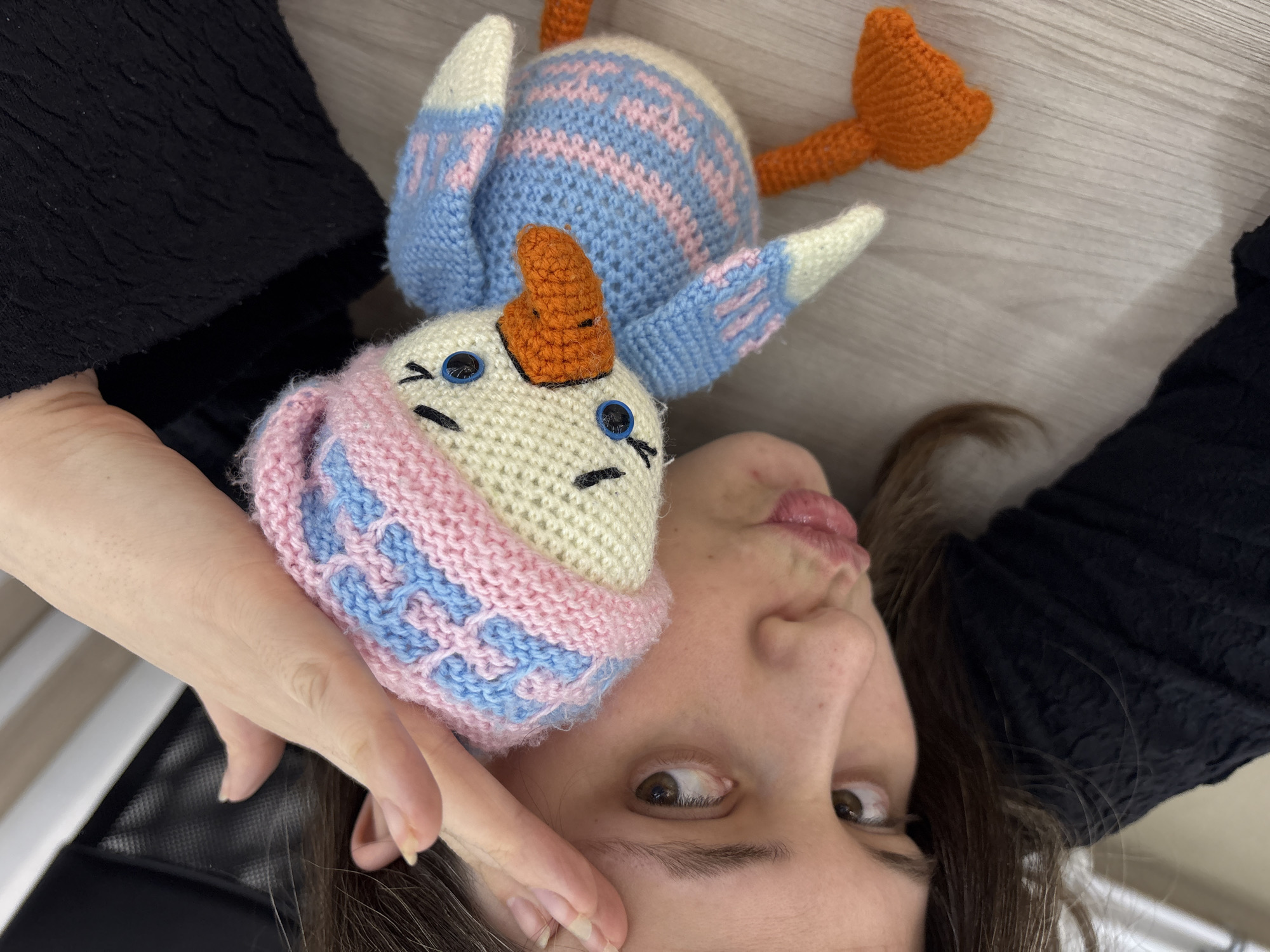 A woman is lying down facing upwards with a knitted duck toy next to her head. The woman has brown eyes and brown hair, and is wearing a black top. The toy duck is made of light-colored yarn with blue and pink detailing, an orange beak, and blue button eyes. The background is a light-colored wooden surface. The location is Chaikovsky, Russia.