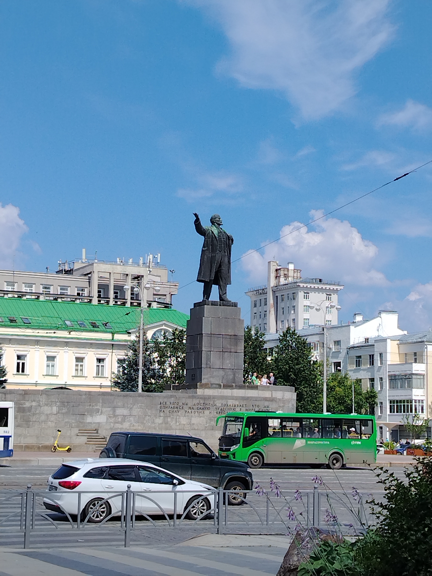A statue of a man in a suit stands on a tall stone pedestal in a city square. The statue is in the center of the frame, with buildings and trees in the background. In front of the statue, there is a green bus with people inside, a dark SUV, and a white station wagon. A yellow scooter is parked near a wall. The sky is blue with some white clouds. The overall scene depicts a public space in a city on a sunny day.