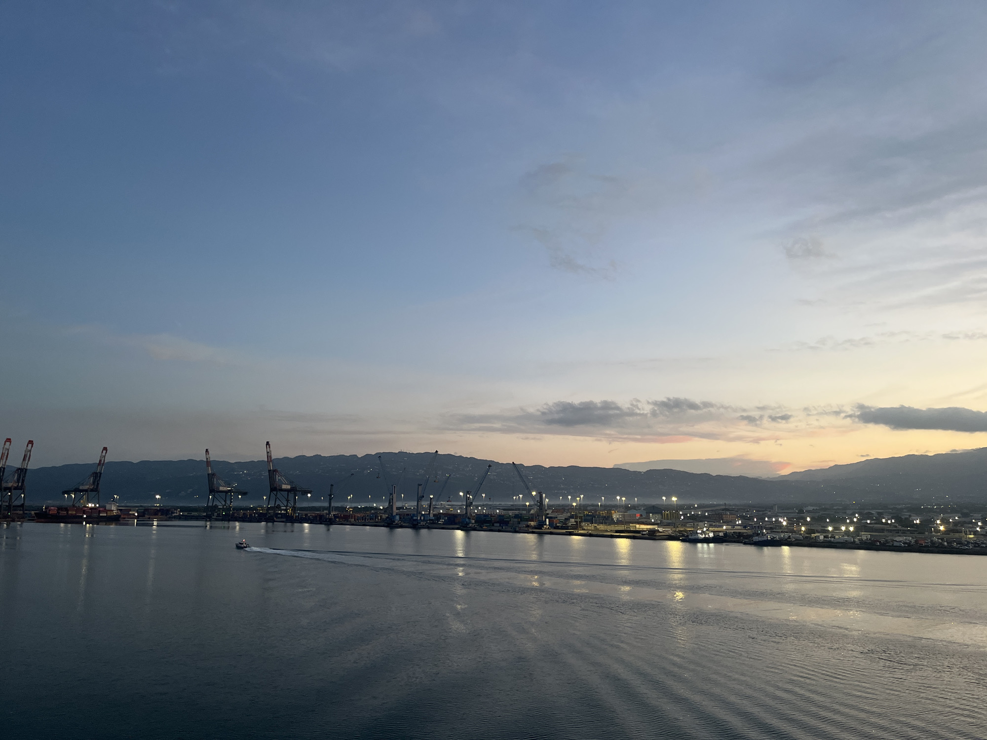 The image depicts a port scene at dusk. In the foreground is a body of water with faint ripples. A small boat is moving across the water, leaving a visible wake.
The midground consists of a port facility with numerous cranes and industrial buildings. The cranes are tall and have a red and white color scheme. Lights illuminate the port area, indicating nighttime activity. Beyond the port, there are hills or low mountains, which form the backdrop.
The sky transitions from a light blue at the top to a yellowish-orange hue near the horizon, suggesting either sunrise or sunset. A few scattered clouds are present. The location is Unknown, Unknown.