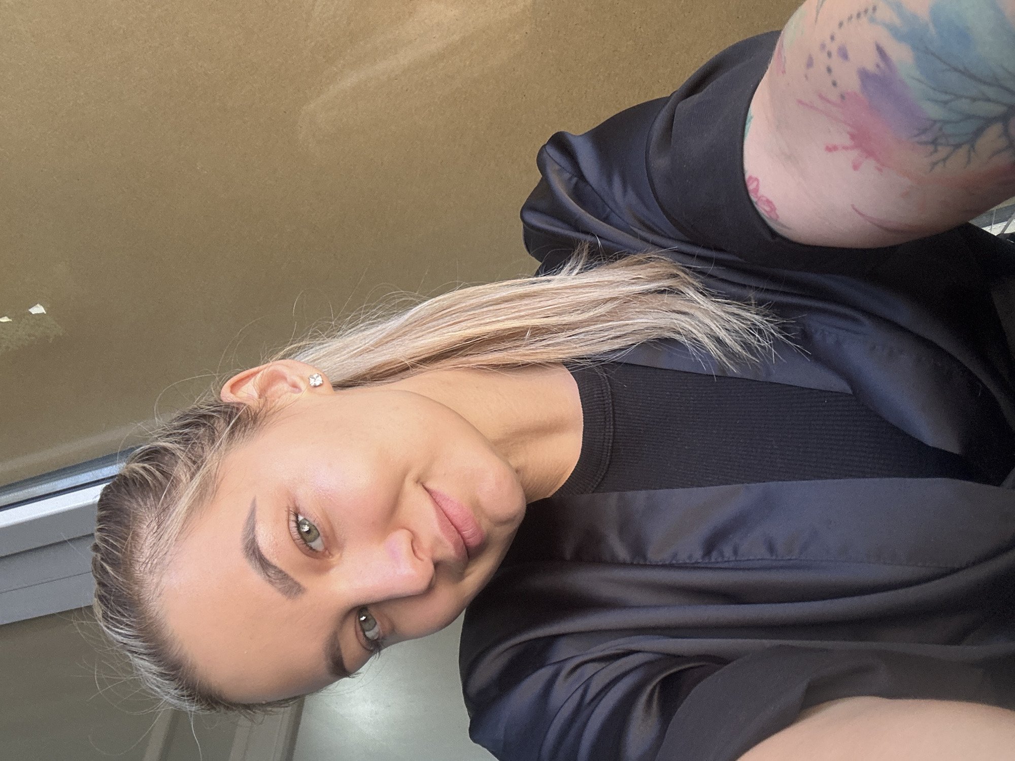 A close-up selfie, captured in Budva, Montenegro, features a woman with blonde hair and striking green eyes, looking directly at the camera with a subtle, pleasant smile. Her head is tilted, and she is positioned diagonally within the frame. Her light blonde hair shows darker roots and appears to be pulled back from her face. A small, square, clear stud earring adorns her right ear. On her visible left upper arm, a vibrant watercolor-style tattoo displays abstract natural or floral motifs in shades of blue, pink, and purple. She is dressed in a black ribbed top beneath a black, satin-like robe or jacket. The background is a plain light brown wall or ceiling, with a white window frame partially visible on the left, indicating an indoor setting illuminated by natural light. There is no visible text in the image.