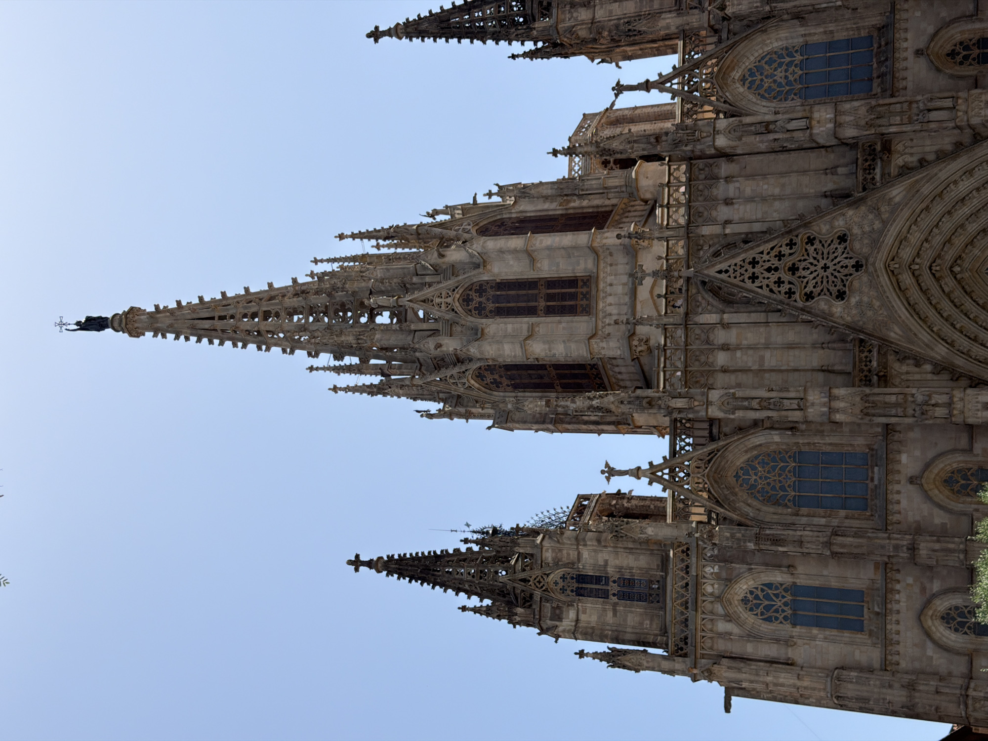 This media file is an eye-level, vertical shot of the exterior of a Gothic cathedral, likely in Barcelona, Spain, given the provided location context. The primary subject is the intricate architecture of the cathedral's spires and facades. Sunlight suggests it is daytime, with clear blue skies indicating fair weather. The ornate stonework features numerous pointed arches, elaborate tracery in the windows, and decorative gargoyles or other sculptural elements typical of Gothic design. The image is dominated by the stone structures, with the sky providing a bright, uncluttered background. There are no people or specific activities visible; the focus is purely on the architectural grandeur. The perspective is angled upwards, emphasizing the height and complexity of the cathedral's design. The overall mood is one of awe at the historical and artistic craftsmanship. No text is visible in the image.