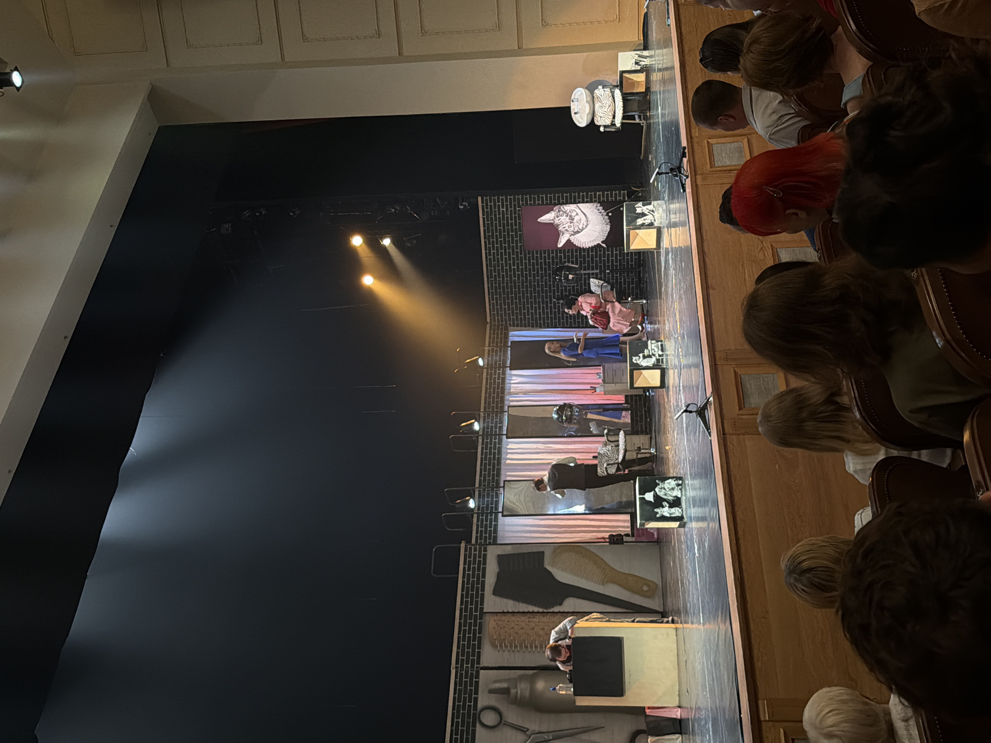 The image captures a live performance on a stage, viewed from the audience. The stage backdrop is decorated with enlarged images of hairdressing tools such as combs, scissors, and spray bottles. Performers are dressed in costumes, and the stage is illuminated with spotlights. A screen displays a graphic of a figure with an elaborate hairdo. Members of the audience are visible, seated in rows of chairs. The scene appears to be taking place at the National Theatre of the Udmurt Republic in Izhevsk, Russia.