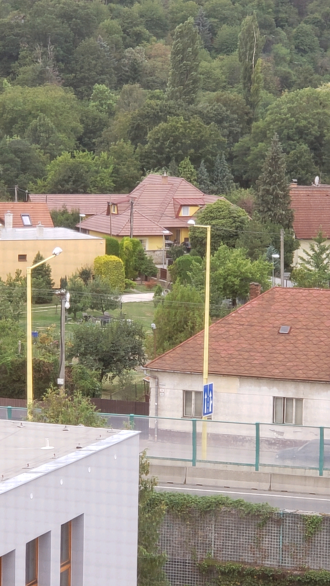 The image depicts a residential area with several houses and a backdrop of dense trees. In the foreground, a modern building with a light grey facade and rectangular windows is partially visible. Adjacent to this, a road with a concrete barrier and a green metal fence runs horizontally. A dark-colored car is partially visible on the road. Further back, several houses with red tile roofs are seen. One yellow house has a prominent gable roof. Two streetlights with yellow poles are positioned along the road. A blue sign with white figures indicating a restroom is mounted on one of the yellow poles. The area is surrounded by lush green trees and vegetation, suggesting a suburban or semi-rural setting. The overall impression is that of a quiet, residential neighborhood.