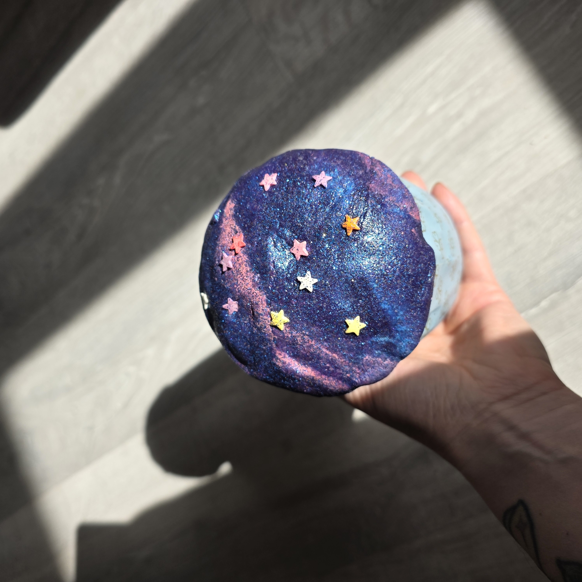 A close-up shot shows a person's hand holding a round object decorated with glitter and star-shaped sprinkles. The object has a blue and purple hue, resembling a cosmic or galactic theme. The background is blurred, but shows a wooden surface with shadows cast across it. The scene is brightly lit with natural light. The location is Unknown, Unknown.