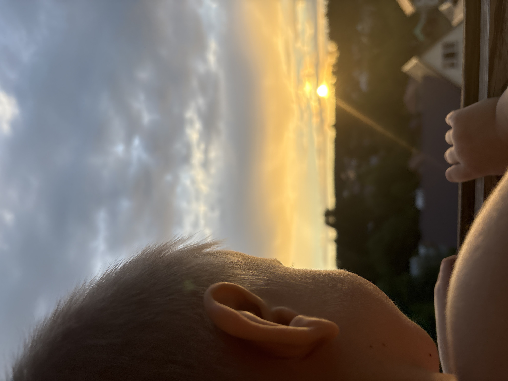 The image captures a close-up, low-angle view of a young child's head and ear against a dramatic sunset sky. The child's fair hair is visible, as is the curve of their ear, suggesting a profile view. The setting is outdoors, with a glimpse of rooftops and trees in the blurred background, indicating a residential area, possibly in Chaikovsky, Russia. The sun is low on the horizon, casting a warm, golden glow that illuminates the clouds and creates a sense of a peaceful evening. The clouds are a mix of grey and bright orange, with the sun's intense light appearing as a bright orb. The overall mood is serene and contemplative, perhaps suggesting a moment of wonder as the child observes the sunset. There is no visible text in the image.