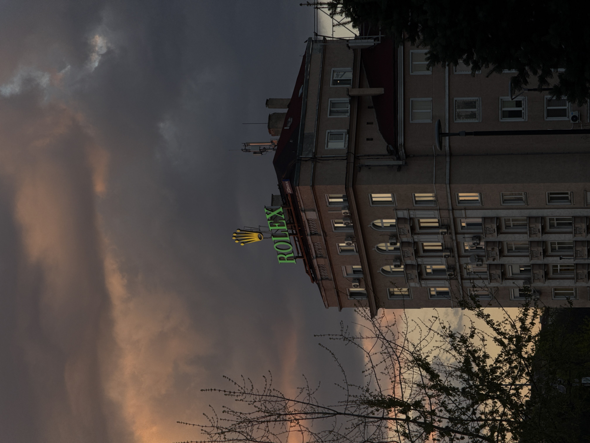 A photograph depicts a building with a large, green and gold "ROLEX" sign affixed to its side. The sky behind the building is filled with dark, dramatic clouds tinged with orange light. In the foreground are bare tree branches silhouetted against the sky. The building is likely located in Sofia, Bulgaria, indicated by the Rolex sign and the architectural style of the building.