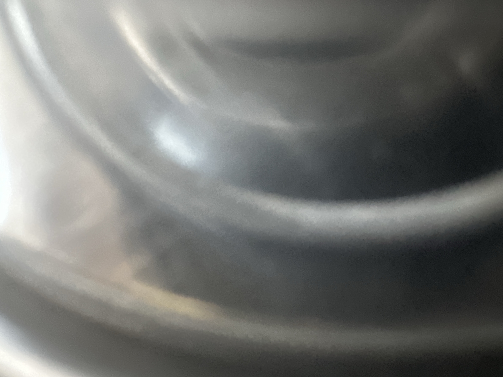 This is a close-up, abstract photograph of a metallic surface, likely stainless steel, with light reflecting off its curved contours. The surface appears smooth and polished, with varying shades of grey, white, and black, suggesting a three-dimensional form. There are no discernible objects, people, or scenes visible. The lighting creates a dynamic interplay of highlights and shadows, giving the impression of movement or depth within the texture. The image is abstract and lacks specific details about time of day, weather, or location cues beyond the provided context of Belgrade, Serbia. There is no visible text in the image.