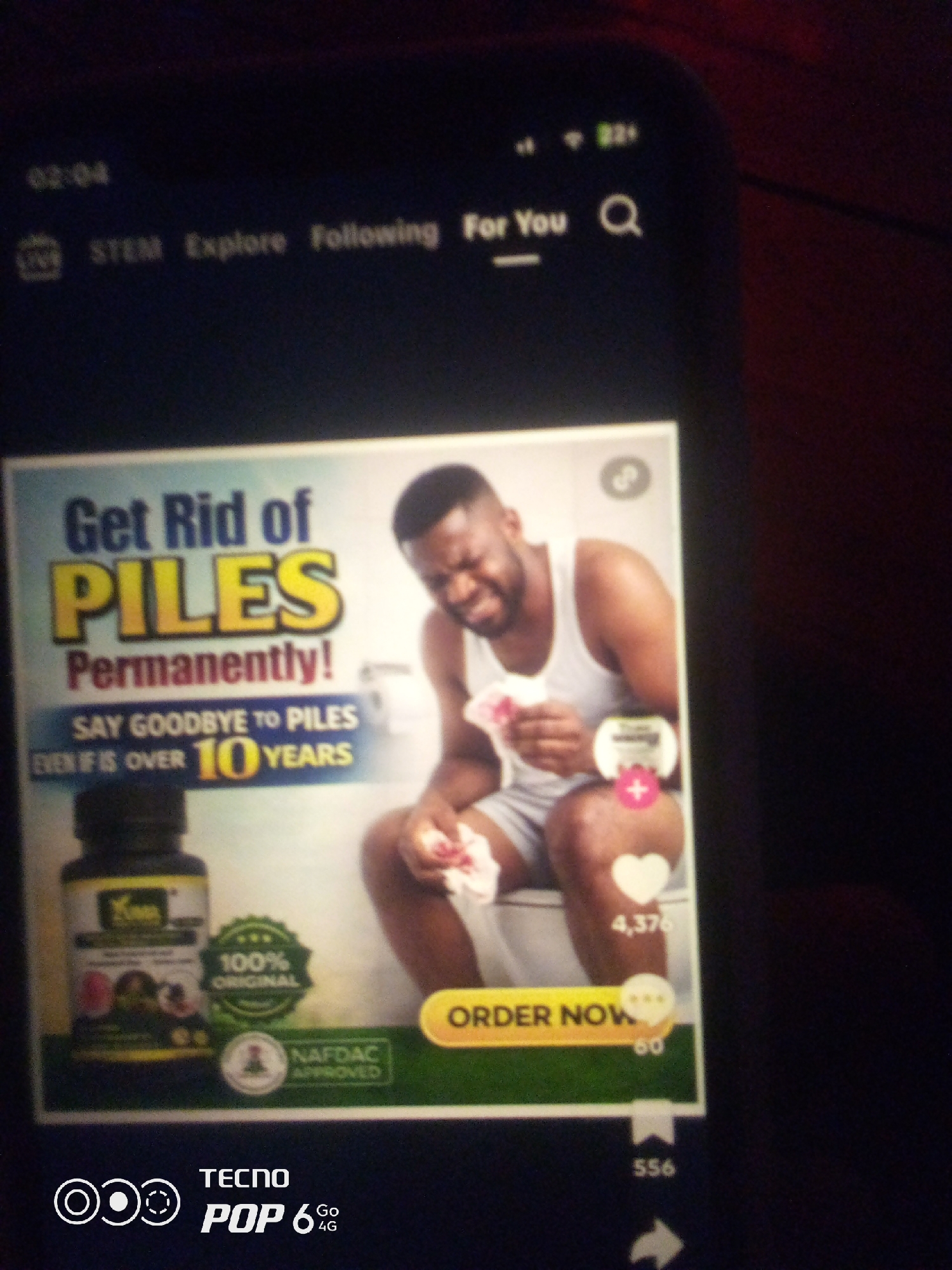 The image displays a mobile phone screen showing an advertisement for a product claiming to "Get Rid of PILES Permanently." The advertisement features a man sitting on a toilet, looking distressed and holding tissue. Text on the screen includes "SAY GOODBYE TO PILES EVEN IF IS OVER 10 YEARS." A bottle of what appears to be herbal medicine is also visible, along with a "100% ORIGINAL" and "NAFDAC APPROVED" stamp. The bottom of the screen shows "TECNO POP 6 4G". The scene is a close-up of the phone screen, with the background being dark and out of focus, suggesting it is in a dimly lit environment, possibly a room. There is no indication of the location being Howayi, Nigeria.