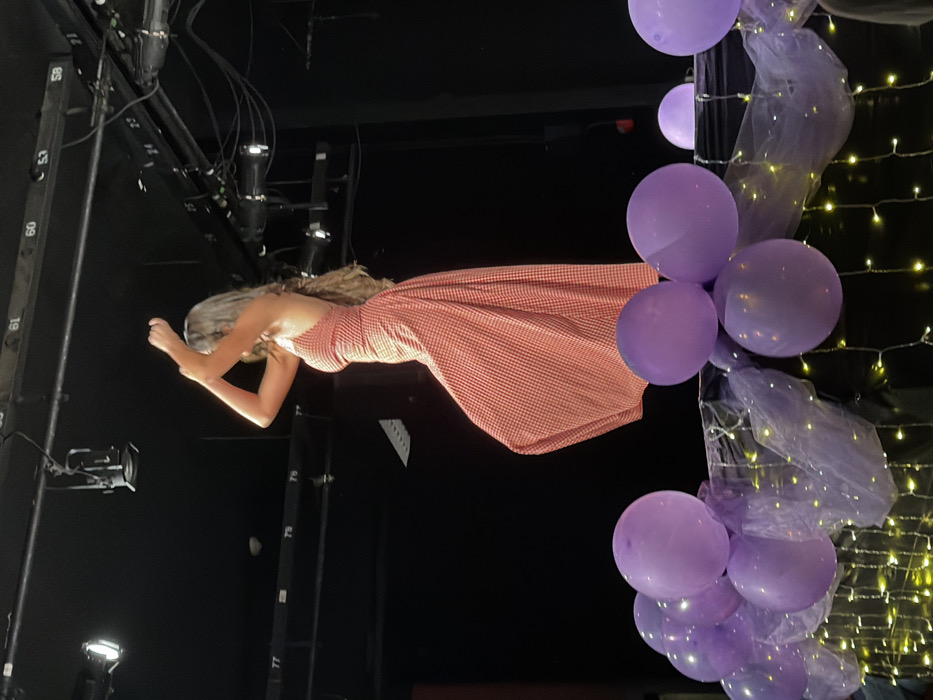 The image depicts a performance setting within an unconfirmed venue in Palos Verdes Estates, United States. A single individual, observed from a side profile, stands center-frame on what appears to be an elevated stage. The individual has long, wavy, light-colored hair extending down their back and is wearing a red and white gingham-patterned maxi dress with a halter neckline and an open back design. Their arms are raised above their head, bent at the elbows, with hands positioned together, suggesting a dynamic pose or dance movement.
In the foreground, a dark structure or railing is decorated with multiple matte purple balloons, purple sheer fabric (tulle), and warm-toned string lights. The balloons partially obstruct the lower portion of the central figure.
The background is dark, revealing a stage environment. Overhead, black structural elements, likely stage rigging, are visible with multiple mounted stage lights or spotlights. White numbers (e.g., "77", "78", "79", "59", "60") are affixed to these structures. The lighting primarily illuminates the central figure and the foreground decorations, with the rest of the background remaining dim.