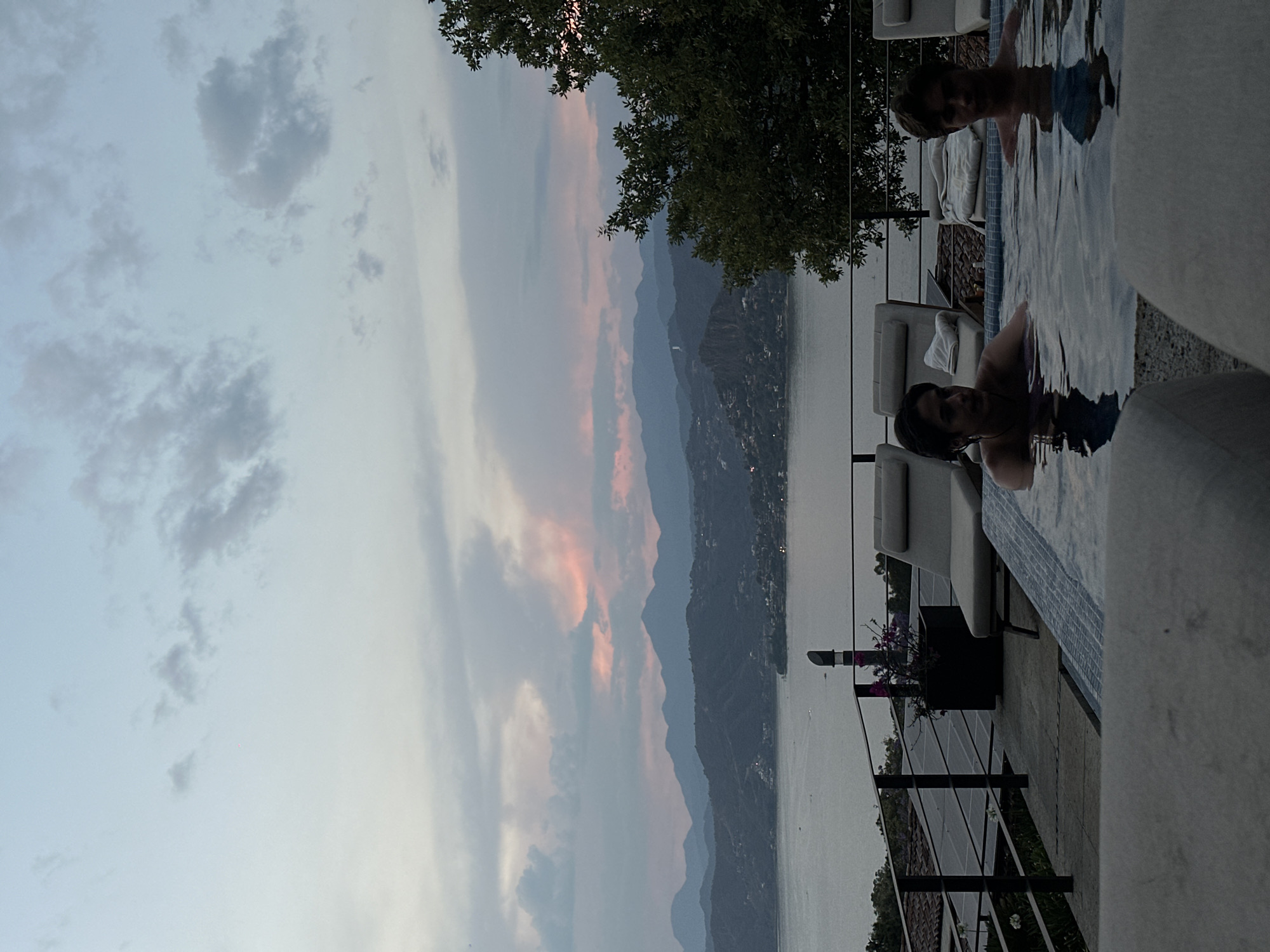 The image shows two people in a swimming pool overlooking a large body of water and mountains. The sky is cloudy with hints of sunset. White lounge chairs are nearby the pool. A black metal railing is visible along the edge of the terrace. The location is possibly a city in an area with mountainous terrain near a large lake or sea, but without additional information, the specific city and region are unknown.