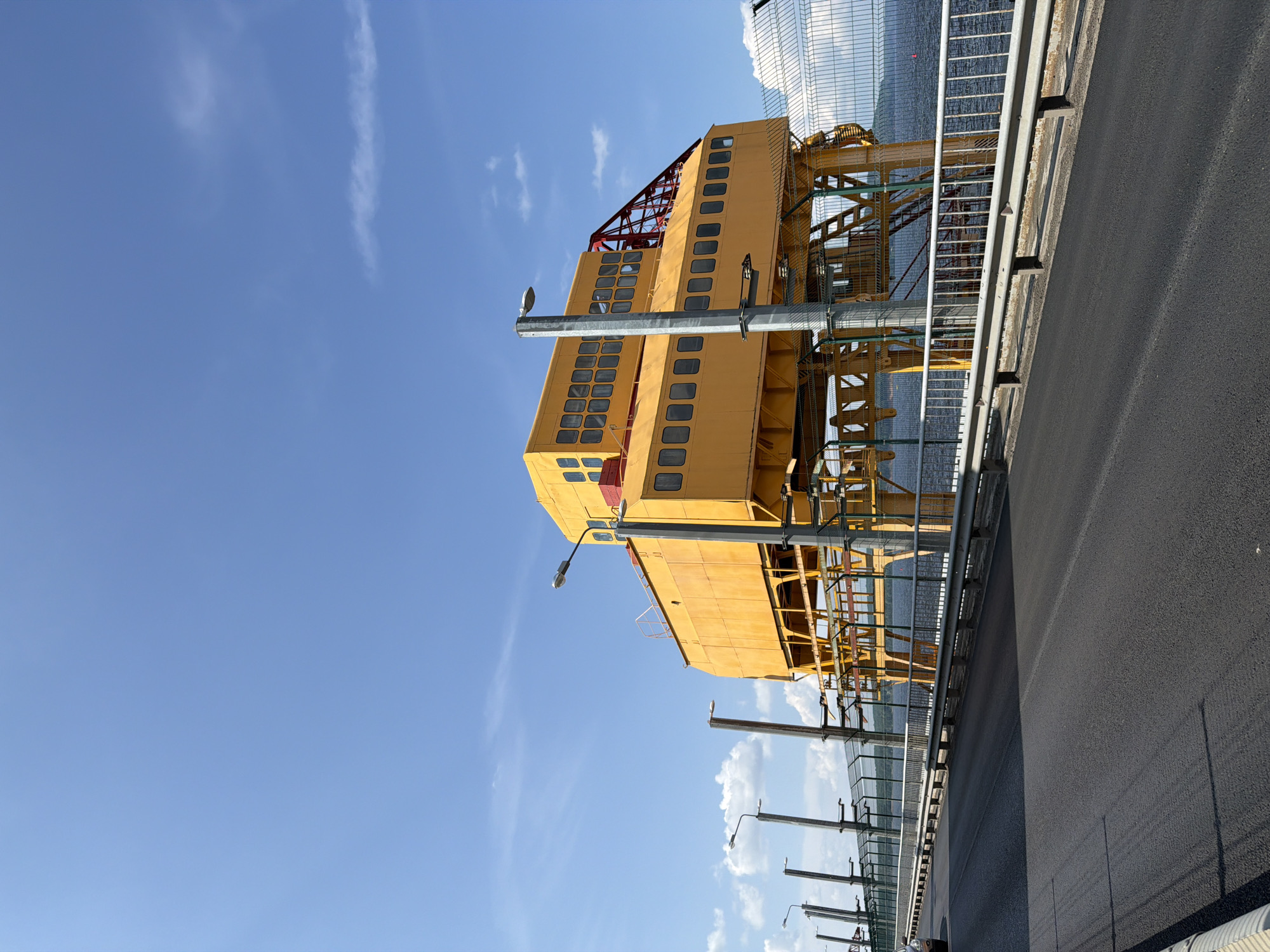 A bright yellow industrial structure is situated on a bridge over a body of water. The structure has multiple windows and is supported by a network of yellow beams and columns. A metal fence runs along the side of the bridge, separating the roadway from the water and the structure. Several lampposts are visible on the bridge. The sky is clear blue with a few wispy clouds. The roadway is paved with asphalt. This structure is likely part of the Chaikovsky Hydroelectric Station in Chaikovsky, Russia.