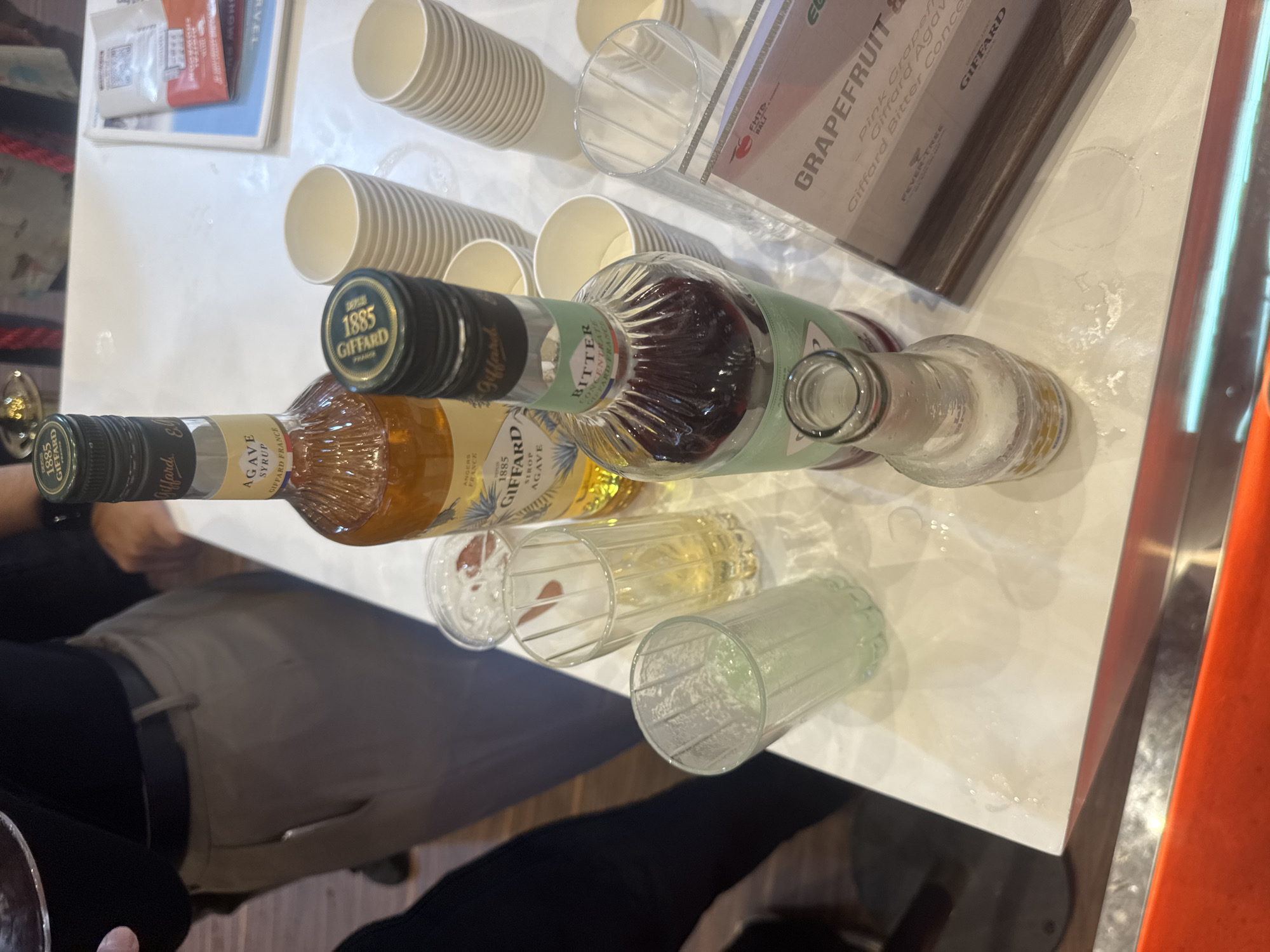 The image captures a tabletop scene, likely at a beverage tasting or promotional event in Kuta Selatan, Indonesia. The main subjects are two bottles of Giffard brand syrups or concentrates, various empty drinking vessels, and part of a person's body.
On a white table surface, two Giffard bottles are prominently displayed. One bottle, with a gold liquid, is labeled "AGAVE SYRUP" and bears "1885 GIFFARD FRANCE" on its cap and label, along with "ANGERS FRANCE." The second bottle, containing a dark red liquid, is labeled "BITTER CONCENTRATE" and also features the "1885 GIFFARD FRANCE" branding. Nearby, there are several stacks of white, disposable paper cups and a few clear, ribbed glasses, some empty and one appearing to contain a light-colored liquid. An empty, frosted glass bottle, possibly a mixer, is also visible.
In the upper right, a display stand with a brown wooden base presents text related to potential drink recipes or ingredients, mentioning "GRAPEFRUIT &," "Pink Grapefruit," "Giffard Agave," and "Giffard Bitter Concentrate," with "FEVER-TREE" and "GIFFARD" also visible. The text "BALI" is partially visible, aligning with the provided location context.
The table surface shows several circular wet marks and small liquid splashes, suggesting active use and recent pouring or mixing of drinks. A person's torso, arm, and hand are visible on the left side of the frame, indicating human presence and interaction with the setup. The lighting suggests an indoor setting. No specific time of day or weather conditions are discernible.