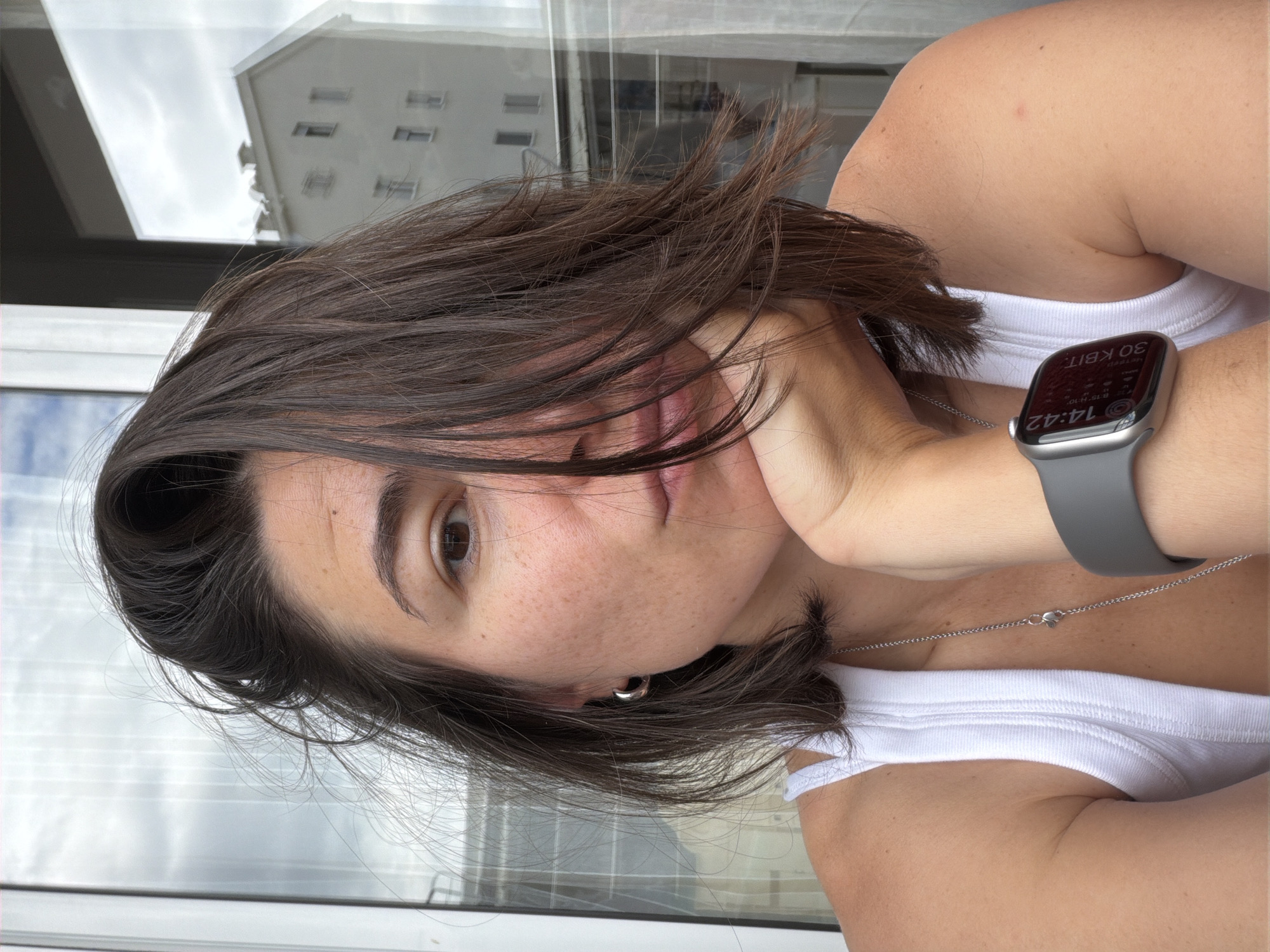 A young adult female is depicted in a close-up, waist-up shot, looking directly at the camera with a neutral expression. She has dark brown hair, brown eyes, and visible freckles across her nose and cheeks. Her left hand is gently resting against her chin. She is wearing a simple white ribbed tank top and a delicate silver chain necklace. On her left wrist, she wears a silver smartwatch with a grey band.
The background shows an outdoor scene, likely viewed from a window or balcony, with a cloudy sky dominating the upper portion. Below the sky, a white building with multiple dark rectangular windows is visible, suggesting an urban or coastal environment. This setting is consistent with the provided location context of Budva, Montenegro.
The activity is a posed photograph. Notable details include the time displayed on the smartwatch screen, which reads "14:42" (2:42 PM). Other visible text on the watch face includes "30 KBIT", "8:15", "H-10", and the "Apple" logo. The overall ambiance is casual and relaxed, captured during what appears to be an overcast day.