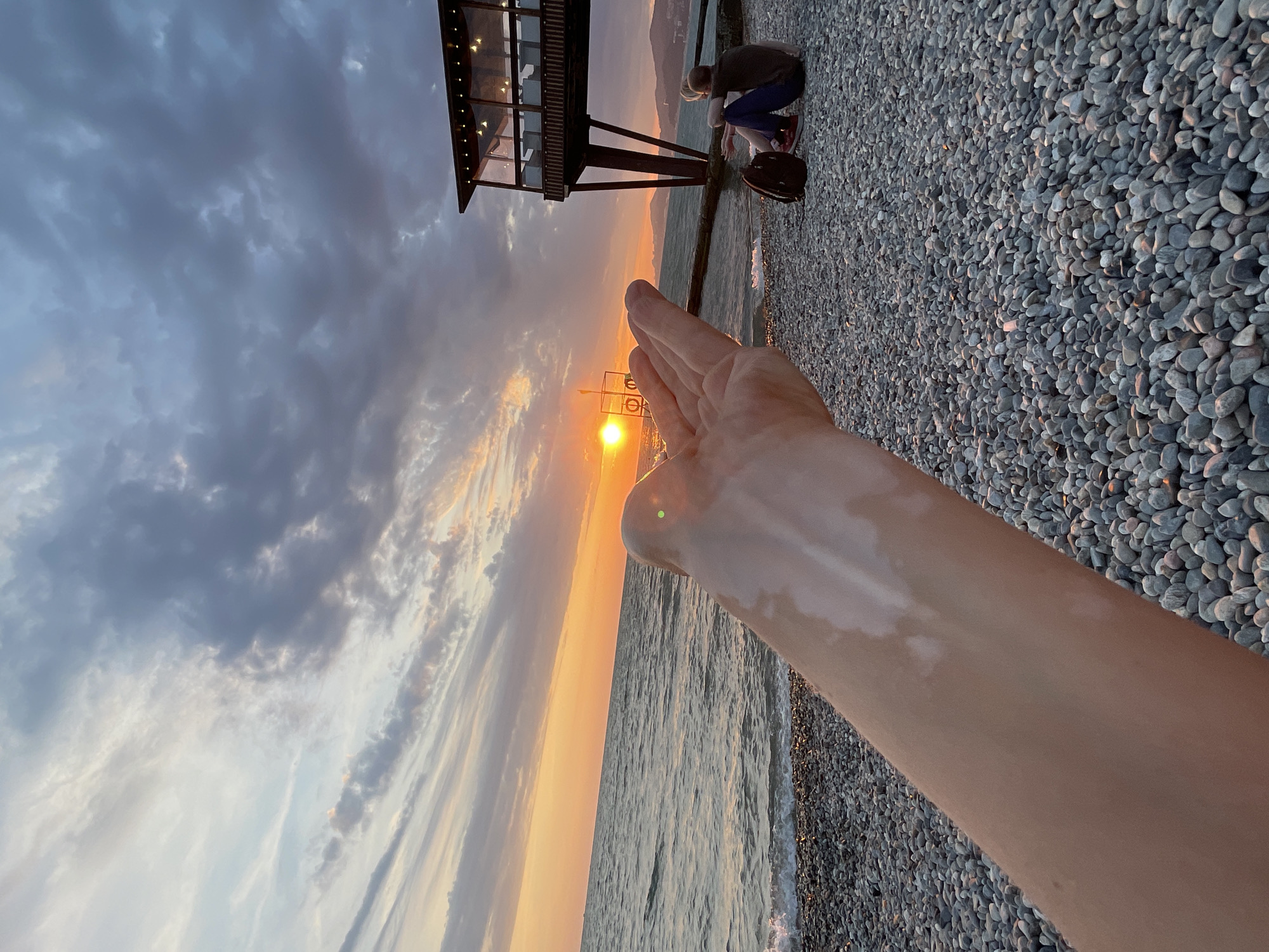 The image depicts a sunset on a pebble beach in Sochi, Russia. In the foreground, a person's hand is extended, palm up, as if to catch the sun. The sun is setting over the sea, casting a warm orange glow on the water and the sky. The sky is filled with dramatic clouds, with streaks of orange and purple. In the background, a couple sits on the beach, facing the sea, with a backpack between them. A wooden structure with glass windows, possibly a cafe or restaurant, is visible on stilts over the water. The beach is covered in smooth, grey pebbles. The overall scene is serene and picturesque, capturing the beauty of a sunset on the Black Sea coast.