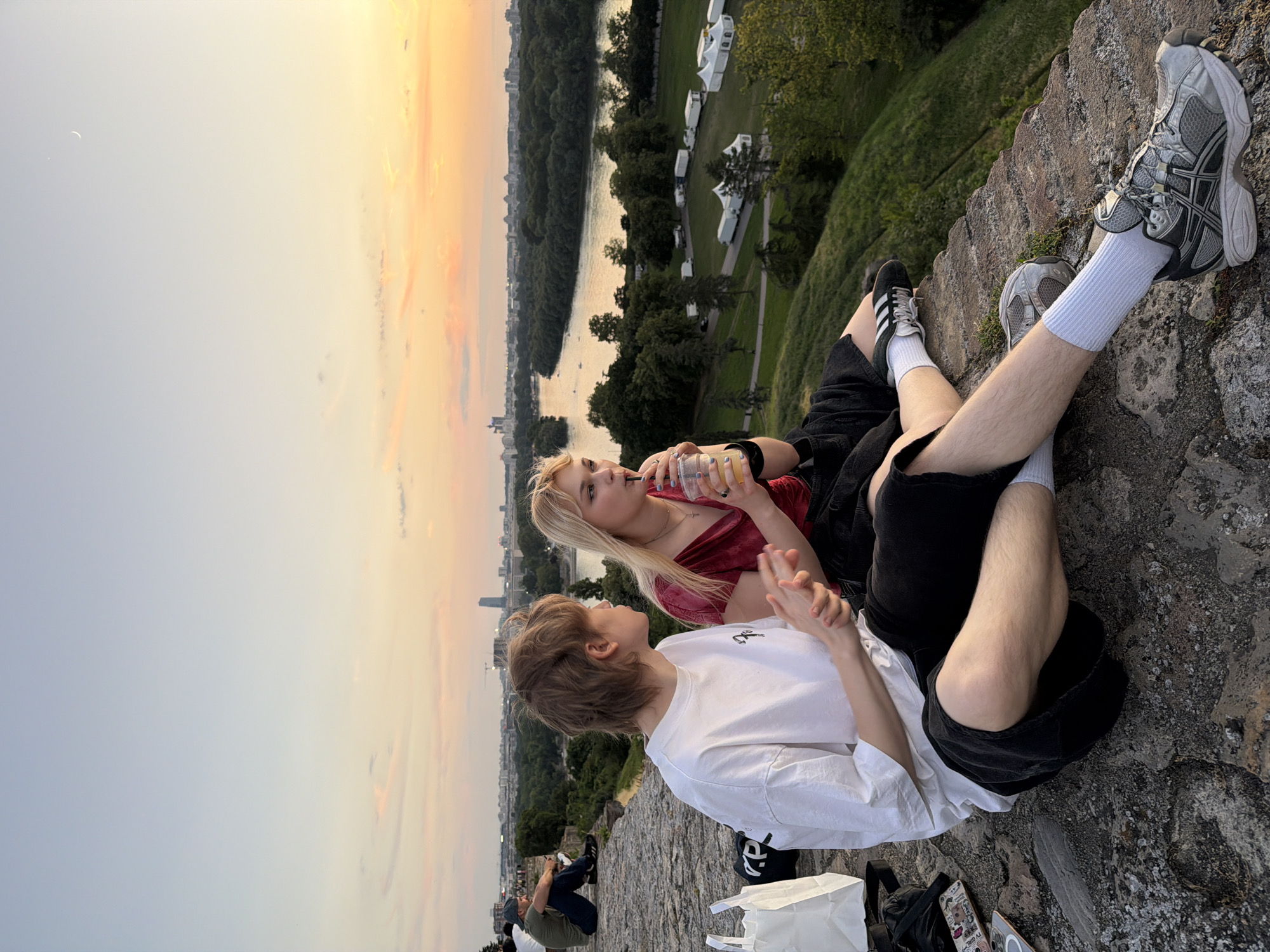 Two individuals, a person with short brown hair wearing a white t-shirt and black shorts, and a person with blonde hair wearing a red v-neck top and black skirt, are sitting on a stone wall overlooking a city and a river at sunset. The person with blonde hair is drinking from a clear plastic cup with a straw. In the background, a river flows through a densely forested area, with buildings and structures visible on the opposite bank and in the distance. A few tents and vehicles are set up in a grassy area below the wall. The sky is a gradient of blue and orange hues, with a crescent moon and a faint star visible. Another person, wearing a cap and dark clothing, is seated further back on the wall. Several bags and a skateboard are placed on the ground near the two main subjects. The overall scene suggests a relaxed outdoor gathering in Belgrade, Serbia, likely at Kalemegdan Fortress due to the prominent view of the confluence of the Sava and Danube rivers and the city skyline.