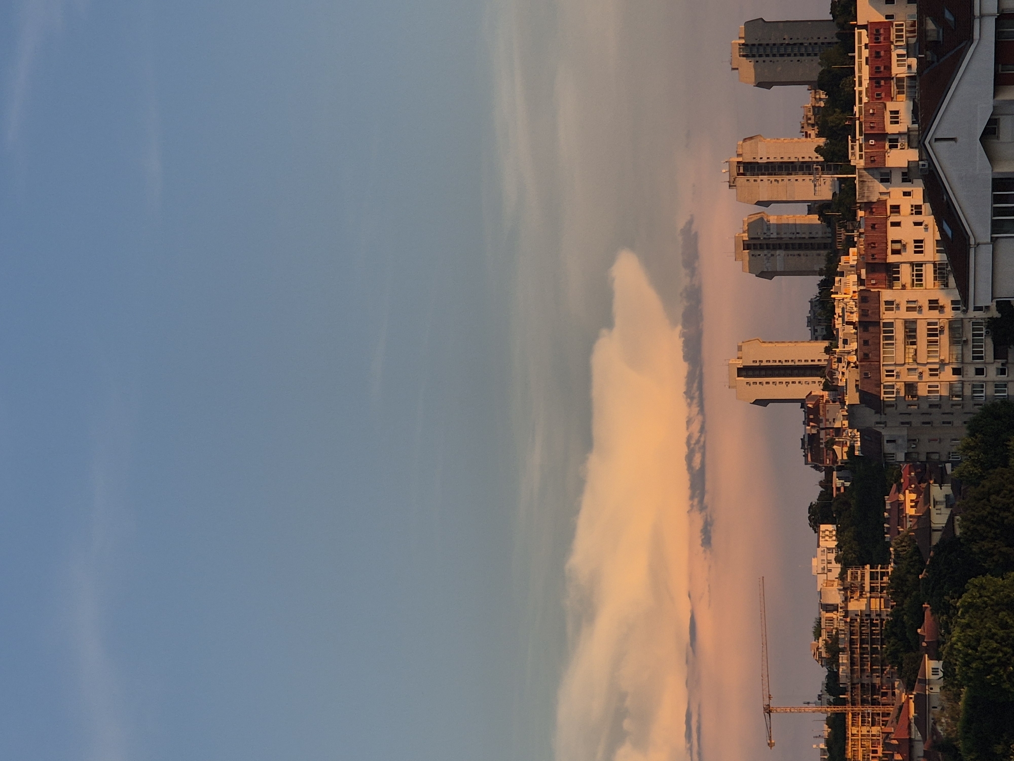 The image captures a cityscape at dusk. The sky transitions from a soft blue at the top to shades of orange and pink near the horizon, illuminated by the setting sun. A large, distinct cloud formation with a mushroom-like cap is visible in the middle of the sky, catching the warm light.
Below the sky, a dense urban landscape unfolds. A construction crane stands prominently on the left side of the image, indicating ongoing development. Various buildings, ranging from multi-story apartment blocks with visible balconies and windows to smaller residential houses with red-tiled roofs, fill the scene. Some buildings appear to be under construction, with visible frameworks and unfinished facades. The architecture suggests a mix of modern high-rises and older residential structures. Trees and greenery are interspersed throughout the city, softening the urban environment. The lighting suggests it is late afternoon or early evening, with shadows starting to form.