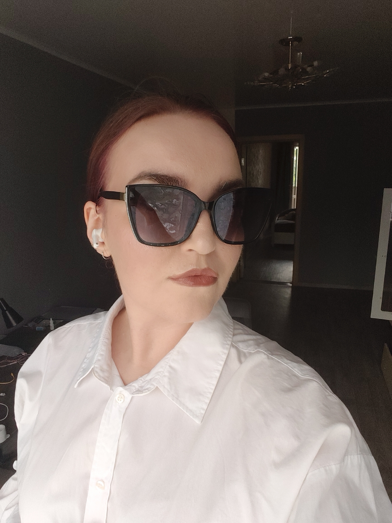 A woman with reddish-brown hair, wearing oversized black cat-eye sunglasses, a white collared shirt, and a white wireless earbud in her left ear, is pictured from the chest up. She is indoors, facing forward, with a slight tilt of her head. The background appears to be a domestic interior, with a dark wall behind her and a doorway visible to her right, leading into another room. A chandelier hangs from the ceiling in the upper right portion of the image. The lighting is dim, suggesting it may be indoors or a twilight setting. The image was taken in Kemerovo, Russia.