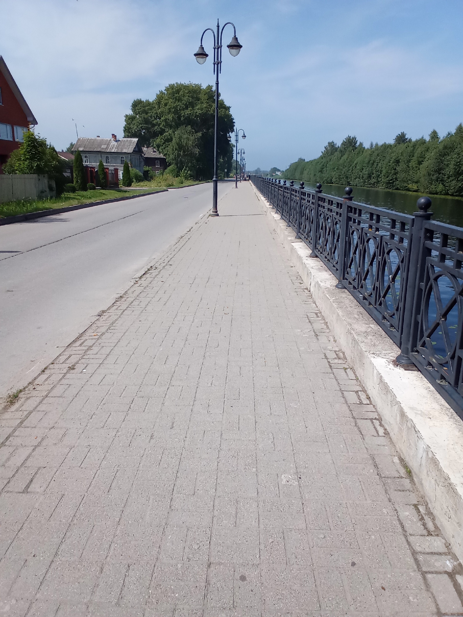 The image shows a sidewalk with a black metal fence along a body of water. The sidewalk is paved with light gray rectangular bricks. The fence has an ornate pattern of circles and lines. Behind the fence, the water is dark and reflects the green trees lining the opposite bank. Across the street from the sidewalk, there are houses and trees. The sky is blue with some scattered clouds. The scene appears to be in Belozersk, Russia, given the architecture and natural surroundings. There are no people visible in the image. The lighting suggests it is daytime, likely late morning or early afternoon.