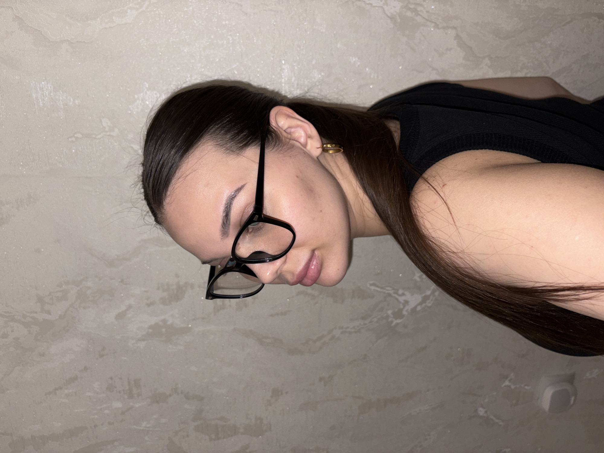 A young woman with dark hair pulled back, wearing black-rimmed glasses and a black top, is seen against a light beige textured wall. She has an earring in her left ear. The scene does not provide any visual indicators to verify its location within Chaikovsky, Russia.