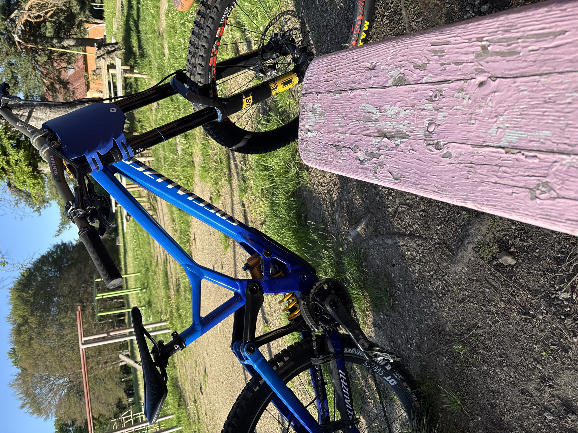 The image shows a blue Specialized Demo mountain bike resting against a pink wooden bench. The bike is in a park-like setting with grass, trees, and playground equipment visible in the background. The location is most likely a park in the city of Samokov, Bulgaria. The bike is positioned with its front wheel near the bench and the rest of the bike extending toward the background. The bench has peeling pink paint. The foreground consists of grass and dirt. The photo is taken outdoors during daylight hours. There are no people visible in the shot.