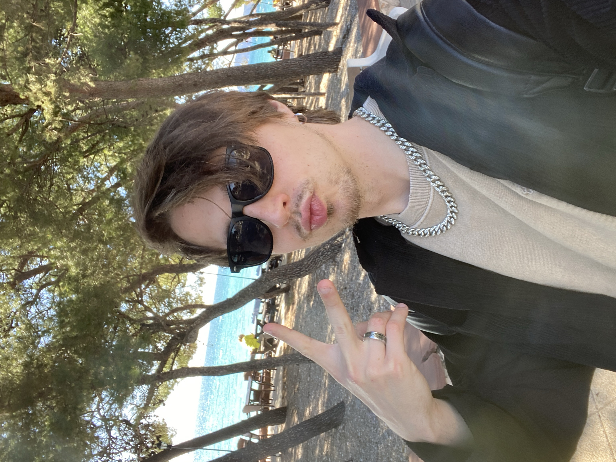 A person is taking a selfie at a coastal location in Bar, Montenegro. The person, wearing sunglasses and a silver chain, is making a peace sign. In the background, the Adriatic Sea is visible, along with trees that provide shade to benches and tables. Other individuals can be seen distantly in the background.