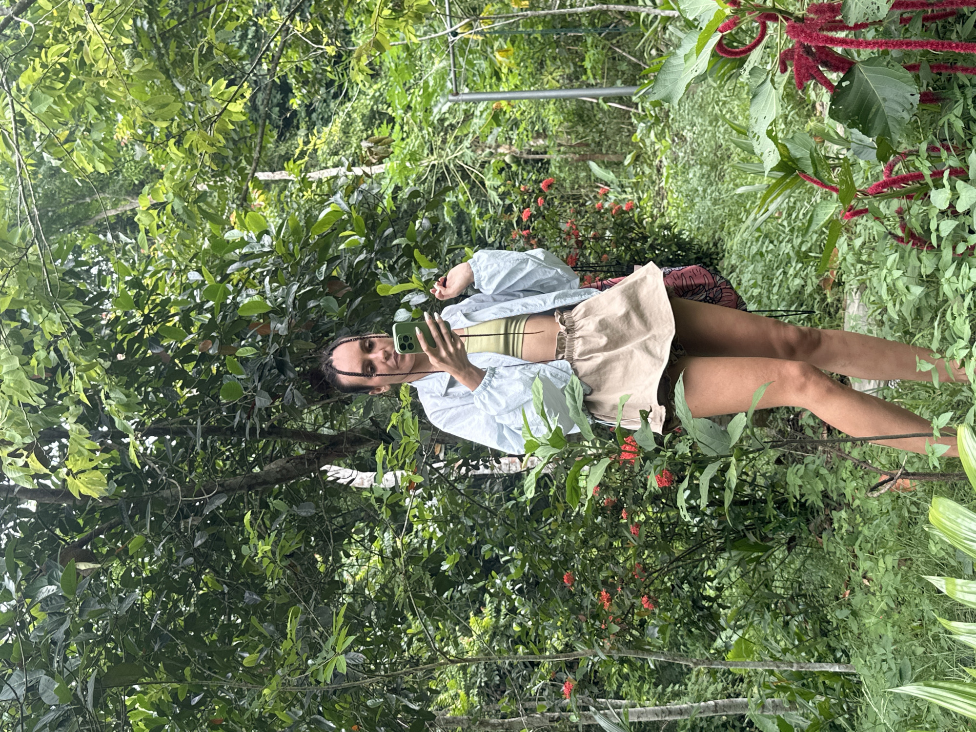 A young woman is standing in a lush, green garden, likely in Cẩm Thạch Commune, Vietnam, taking a selfie with her phone. She is wearing a light blue jacket over a yellow crop top and a beige skirt. The garden is dense with various plants, including trees with large leaves, flowering bushes with red blossoms, and some taller plants with bright red, fuzzy flowers in the foreground. The lighting suggests it is daytime, with natural light filtering through the foliage. The woman is holding her phone in her right hand, angled to capture her reflection in the screen, and her left hand is raised slightly, with her index finger extended. Her hair is styled in two long braids. The overall impression is one of a person enjoying a moment in a vibrant, natural setting.