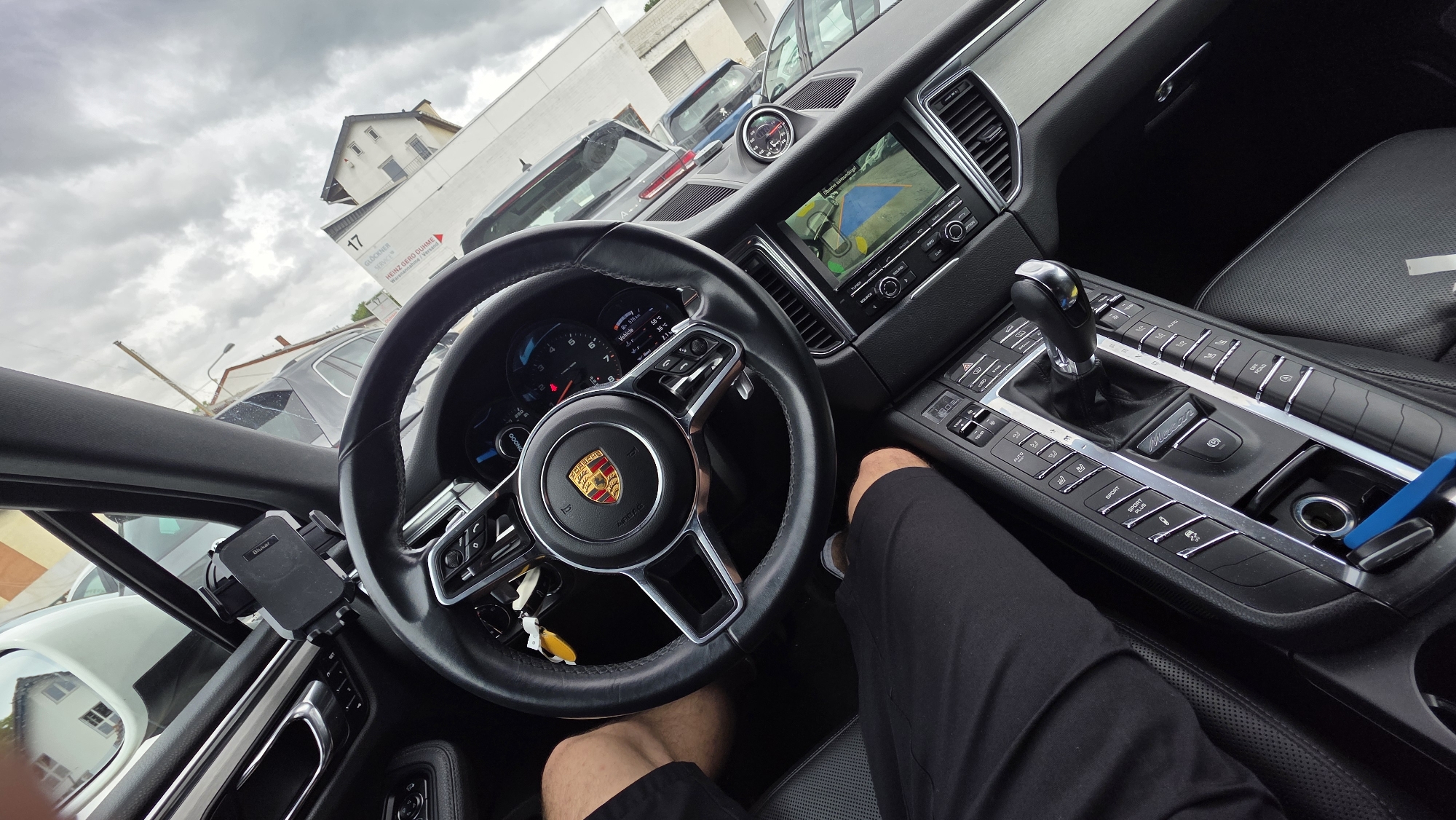 The image shows the interior of a Porsche car, viewed from the driver's seat. The steering wheel, dashboard with a central infotainment screen displaying a parking assist camera view, and the gear shift are prominent. The driver's legs, wearing black shorts, are visible, indicating a person is seated in the driver's position. A phone mount is attached to the dashboard. Outside the car, through the windows, a cloudy sky and a building with the number "17" and text "GLOCKNER SERVICE" are visible, suggesting an urban environment, possibly a car dealership or service center. The image is taken from a high angle, looking down towards the steering wheel and center console.