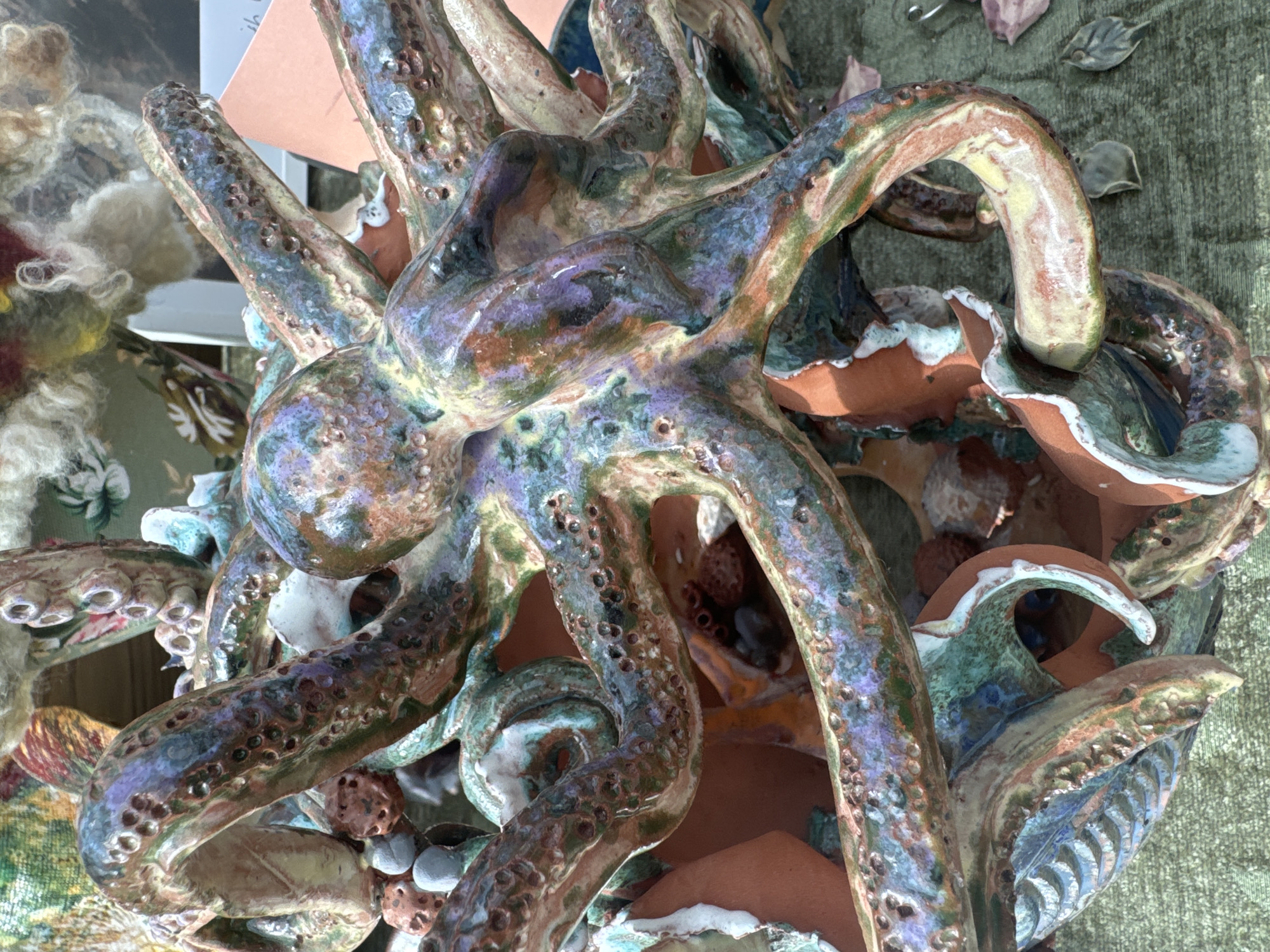 The image shows a close-up of a colorful, multi-tentacled ceramic sculpture resembling an octopus. The sculpture is made of glazed ceramic, with prominent textures and variations in color, including shades of green, purple, blue, orange, and yellow. The tentacles are intricately detailed with small circular indentations, possibly representing suckers. The sculpture is set against a textured green surface, likely a fabric or tablecloth, and in the background, there are blurred elements that suggest a market stall or craft fair, with glimpses of other crafts and decorative items. One of the crops shows a pink card and part of some text, and another shows a woven or felted material. The lighting appears to be natural, suggesting an outdoor or well-lit indoor setting. There are no people or discernible actions visible in the image. The overall impression is of a detailed, artistic craft item. The location is not specified in the image, and therefore, the mention of Kilkenny, Ireland, cannot be verified.