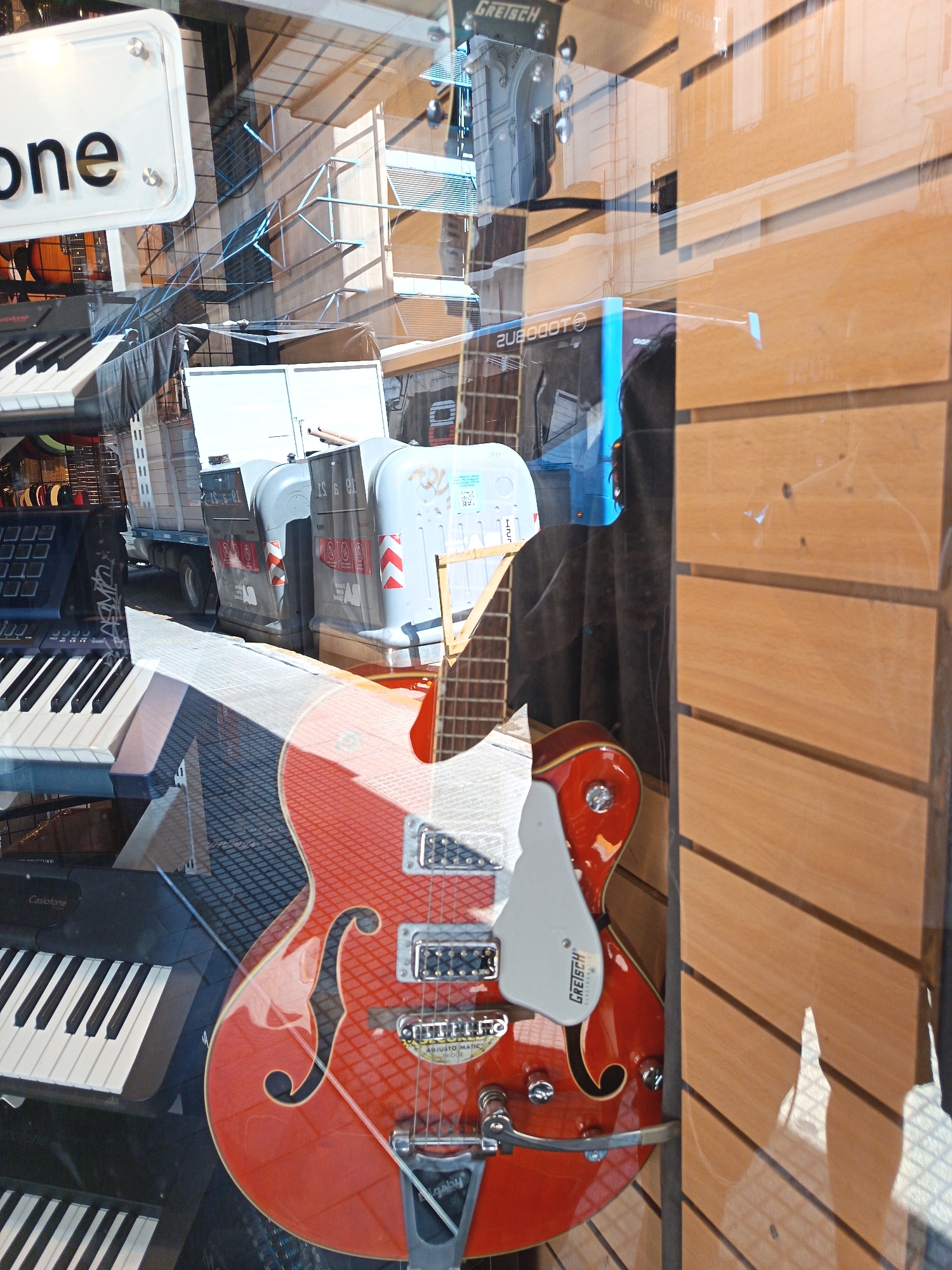 The image depicts a shop window display in Buenos Aires, Argentina. The window reflects the street scene, including scaffolding and a utility truck with large containers. Inside the shop, a red Gretsch guitar is prominently displayed, alongside two keyboards (one Casio) and other musical equipment. A sign with the word "one" is visible. The reflection in the window shows part of a building with architectural details. The shop's interior includes wooden paneling.