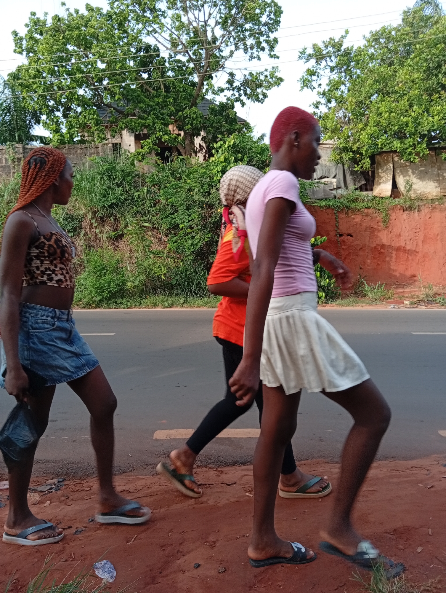 Three young women are walking along the roadside in Nkpor, Nigeria. The woman on the left has braided brown hair and is wearing a leopard print crop top and a denim skirt. She is carrying a black bag. The woman in the middle is wearing a headscarf, an orange shirt, and black pants. The woman on the right has short red hair and is wearing a light pink t-shirt and a white skirt. All three are wearing sandals. They are walking along a road with a red dirt shoulder. The background includes lush green trees and a wall.