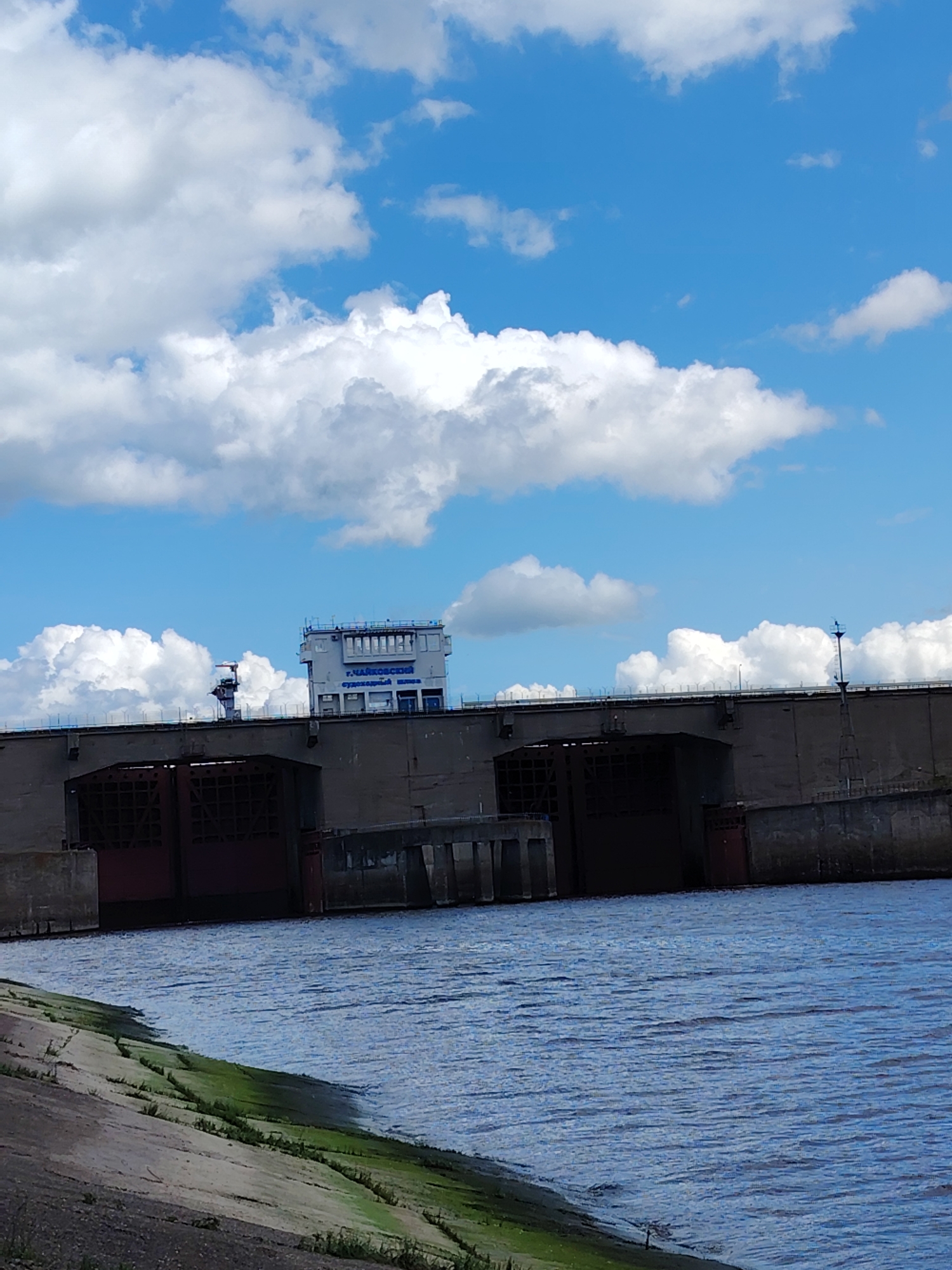 The image depicts a river scene in Chaikovsky, Russia. In the background is the Chaikovsky Dam, a large, concrete structure with several arched openings. A control building sits atop the dam. In the foreground is a body of water with a concrete bank covered in some green algae. The sky above is blue with white clouds. There are no people visible in the image.