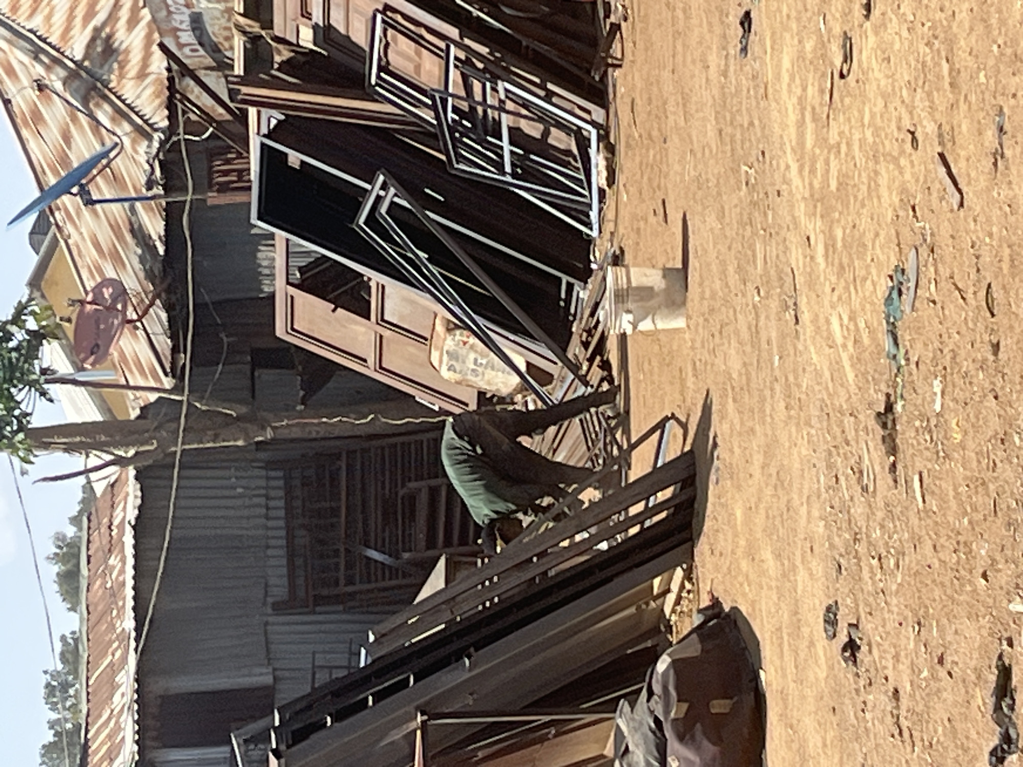 Location: Barakin Akawo, Nigeria. The scene shows an outdoor workshop or storage area, likely involved in metalwork. A man is bent over, working on metal frames or structures. These metal items are stacked around him, possibly doors or window frames. The surroundings consist of corrugated metal structures, suggesting informal or low-cost buildings. The ground is dirt and debris-strewn. There are satellite dishes on the roof.