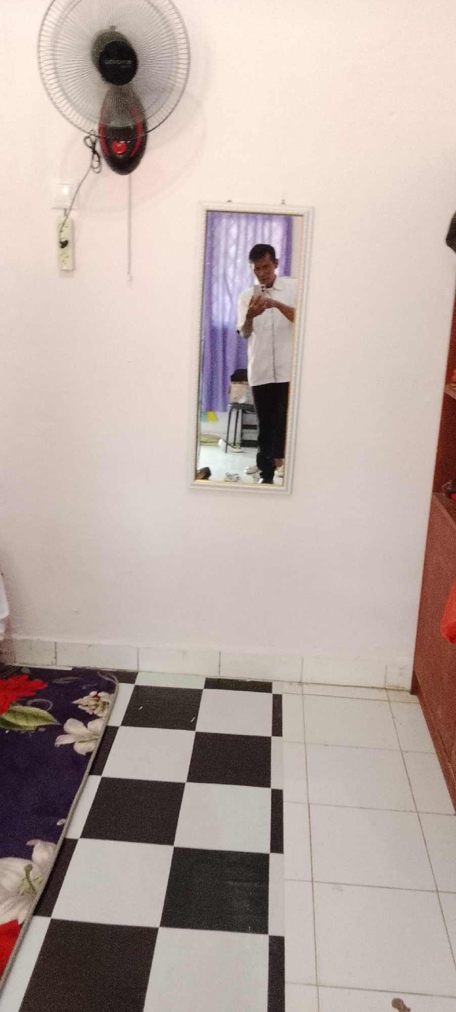 A full-body, vertical mirror selfie is captured indoors, reflecting a man standing in a room. The man is wearing a white button-down shirt and black pants. He is holding a smartphone in his right hand, using it to take the photo. Behind him, a purple curtain is visible, and to the left, a small stool with a black seat and metal legs is partially visible.
The room has a light pink wall and a tiled floor. The floor features a checkerboard pattern of black and white tiles in the foreground, transitioning to plain white tiles further back. A portion of a dark-colored rug with a floral pattern is visible on the left side of the frame, partially covering the black and white tiles.
Mounted high on the pink wall, to the left of the mirror, is a black and red oscillating fan. Below the fan, a white electrical outlet is mounted on the wall. To the far right of the image, the edge of a dark brown wooden cabinet is visible. The overall impression is that of a simple, lived-in interior space. The presence of the fan, rug, and basic furniture suggests a residential or dormitory-like setting. The lighting appears to be ambient, possibly from the room itself or natural light from outside the frame. There is no indication of the specific location within Jepara, Indonesia, beyond it being an indoor environment.
