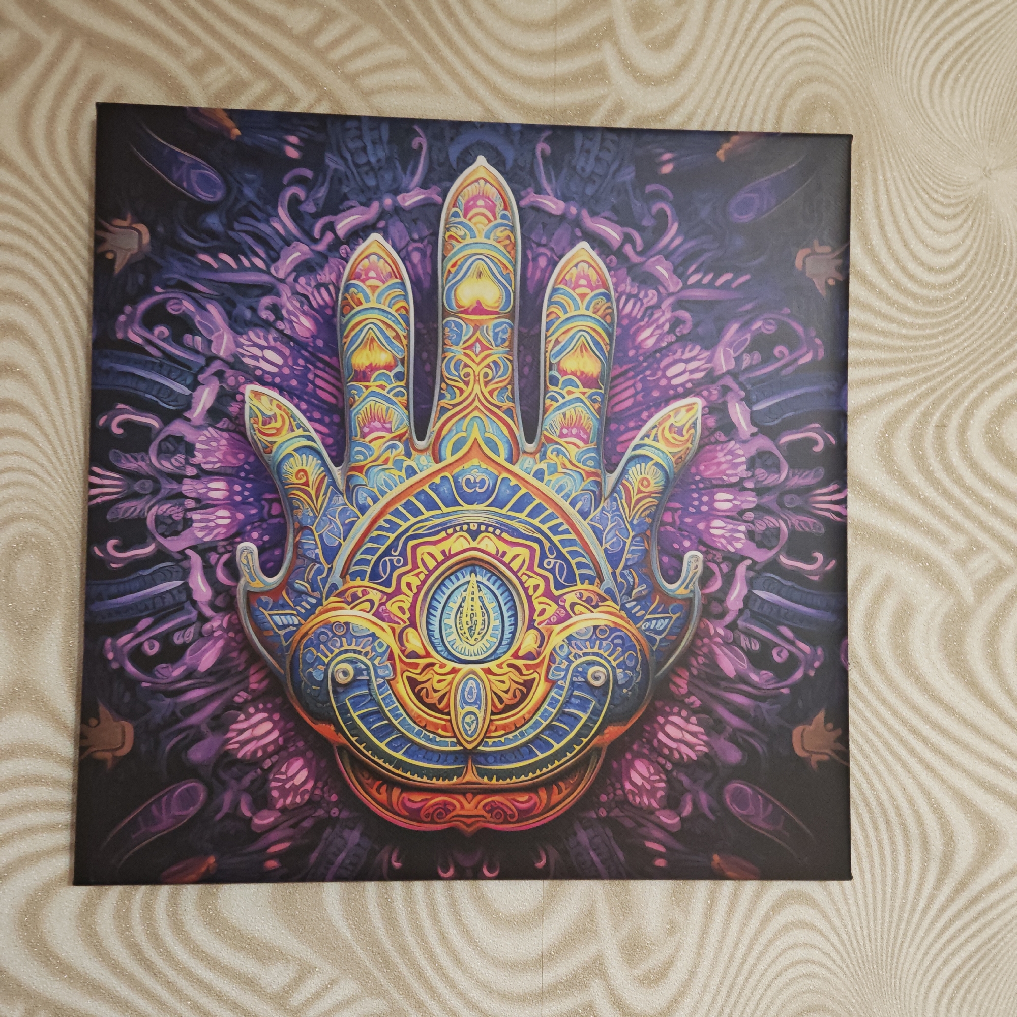 The image shows a square canvas print hanging on a wall with a swirling beige and white wallpaper. The artwork on the canvas depicts a stylized, ornamental hand, possibly a Hamsa, rendered in vibrant colors, primarily blues, yellows, oranges, and purples. Intricate patterns and designs fill the hand and the background, creating a psychedelic or spiritual aesthetic. The hand has a central "eye" design within the palm area. The scene does not appear to be located in Rostov, Russia, as there are no indications of a specific location. The image focuses solely on the artwork and its immediate surroundings.