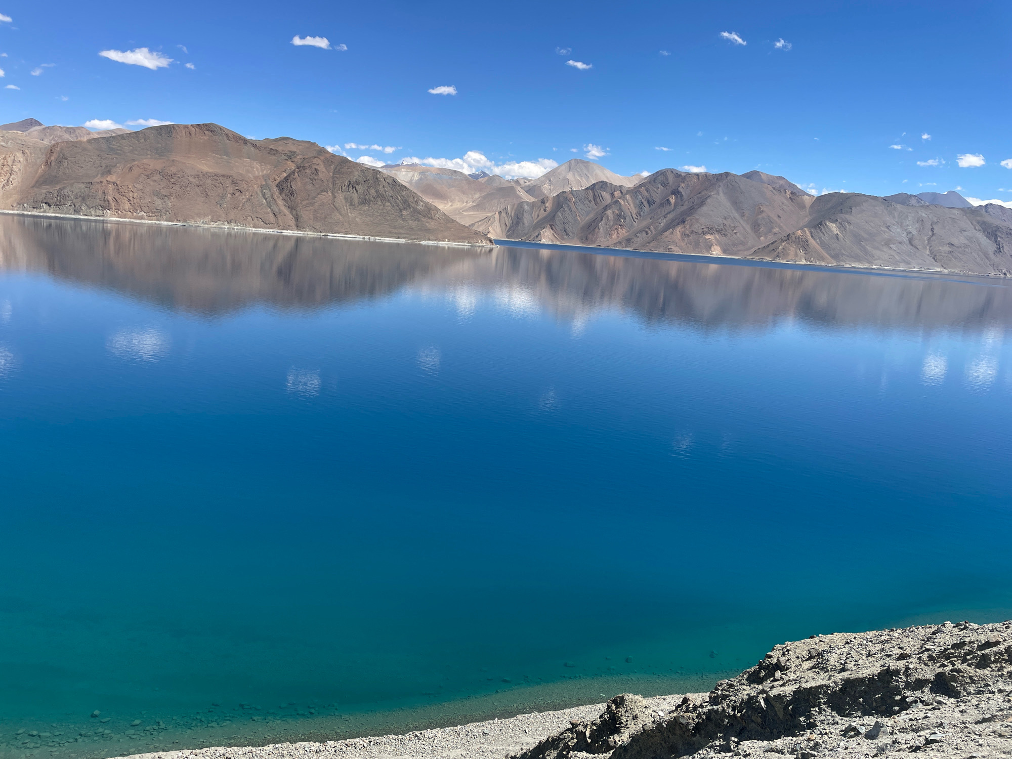 The image displays a vast, calm lake with stunningly clear, turquoise and deep blue waters. The lake is surrounded by rugged, barren mountains that exhibit a range of earthy tones, from light brown to dark gray, with some patches of lighter, more sandy-colored rock. The mountains rise steeply from the lake's edge, and their imposing forms are perfectly reflected on the glassy surface of the water. The sky above is a vibrant, clear blue, punctuated by a few small, wispy white clouds. In the foreground, a rocky, gravelly shoreline is visible, with some larger stones and debris scattered across it. The overall scene is one of natural grandeur and tranquility, characteristic of a high-altitude, mountainous region. Based on the visual cues of the landscape, the location is likely Pangong Tso lake in Ladakh, India.