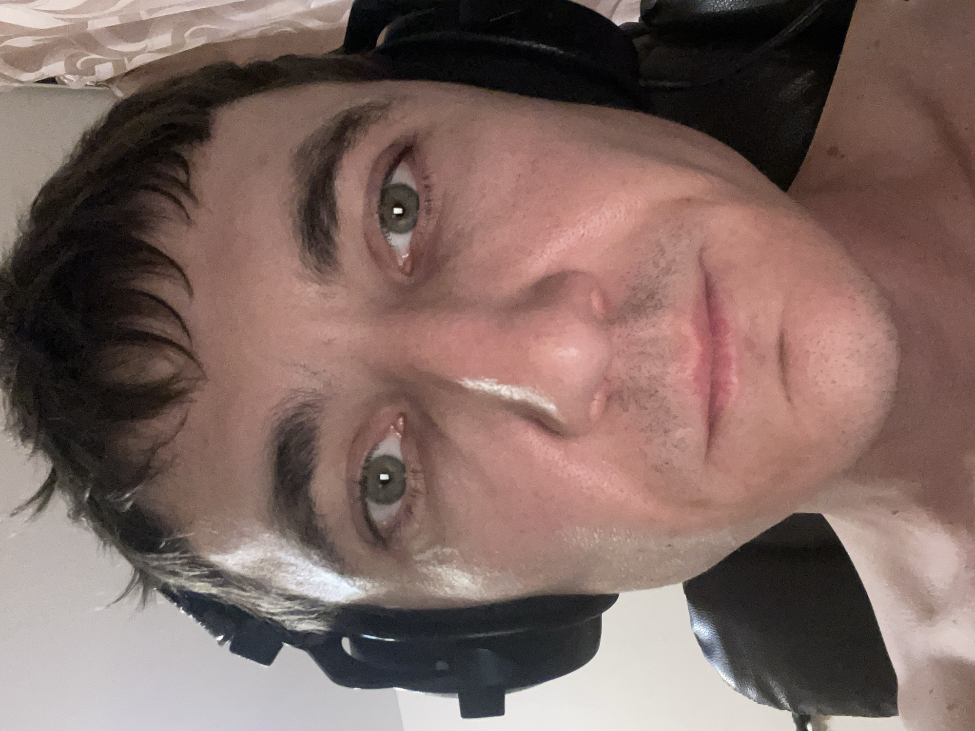 The media file is a close-up, sideways portrait of a person wearing headphones. The subject's face is partially visible, with their eyes and forehead being the most prominent features. They have green eyes and dark hair that appears somewhat disheveled. There is a white mark on their nose, possibly from a product or a smudge. The lighting suggests an indoor setting, and the background is blurry, with hints of fabric and a dark surface, likely furniture. The overall mood is casual and candid, with the subject seemingly looking directly at the camera. No text is visible in the image. The location context provided is Laishevo, Russia.