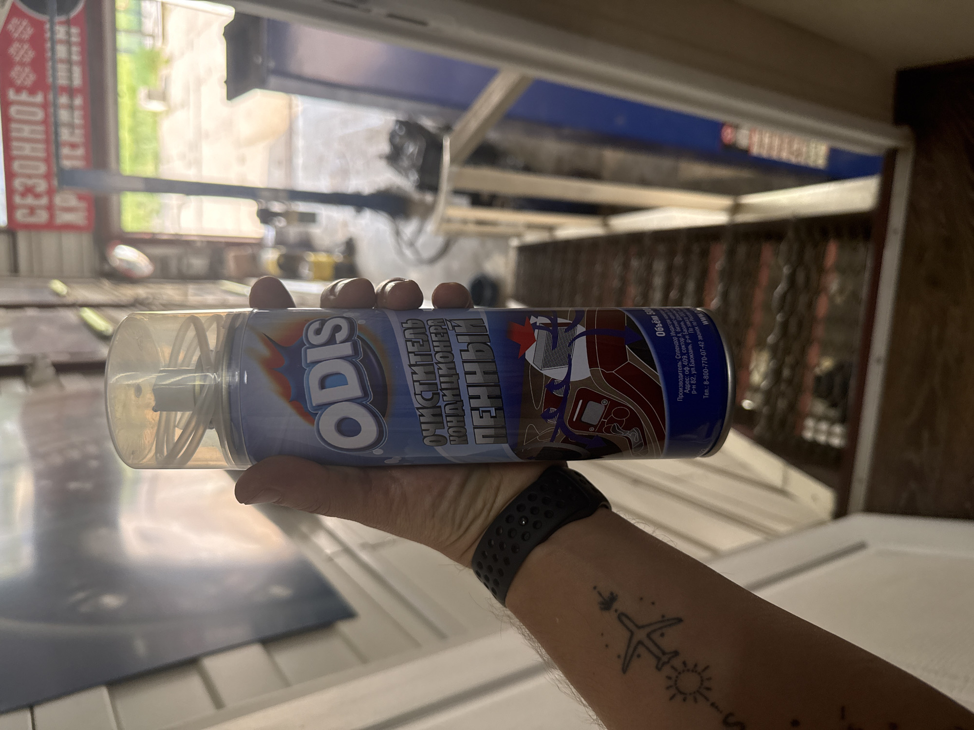 A hand is holding a can of ODIS brand foam air conditioner cleaner. The product is in Russian. The can has a clear plastic nozzle. The hand has a black smartwatch and a tattoo of an airplane, sun, and other symbols. The background is blurry but appears to be inside a garage or workshop, with a window showing greenery outside. The can lists the manufacturer as Splendor Indastri kom, with an address in sector A of a business center in Baoyuan Street, Barany district, Russia. A sign with the Cyrillic text "СЕЗОННОЕ XP" is partially visible.