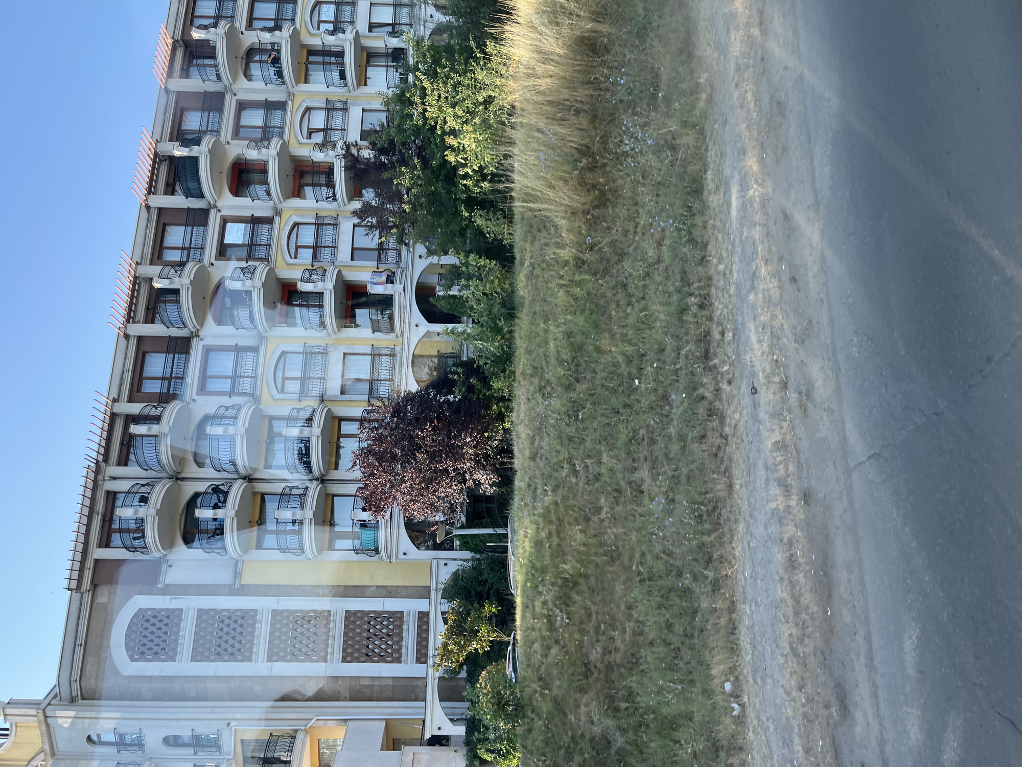 The image depicts a multi-story building with a light-colored facade, adorned with yellow accents and arched balconies featuring black metal railings. The balconies have white curved bases and are topped with decorative wooden pergolas. The building appears to be a hotel or apartment complex. In front of the building, there is a strip of dry grass and a paved road in the foreground. The sky is clear and blue. The location is identified as Nesebar, Bulgaria.