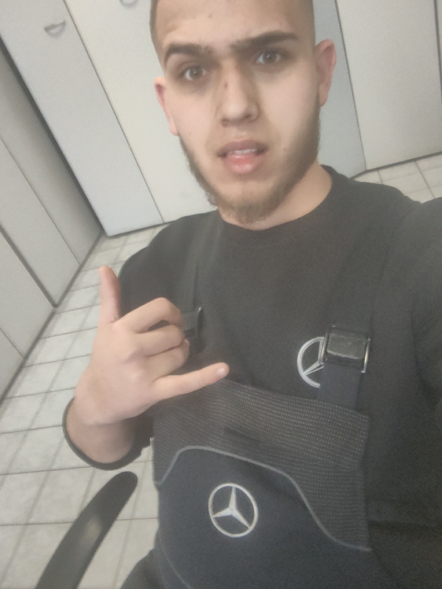 A person with light brown skin, short dark hair, and a light beard is making a "hang loose" hand gesture. They are wearing a dark long-sleeved shirt and a dark-colored chest bag with the Mercedes-Benz logo on it. The background consists of white tiled walls and floor. The location is likely indoors, potentially in Sofia, Bulgaria.